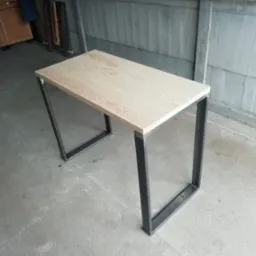 Stół z blatem z jasnego drewna i nogami z profili stalowych o kwadratowym przekroju, spawanych w kształt litery U, surowe otoczenie warsztatowe.