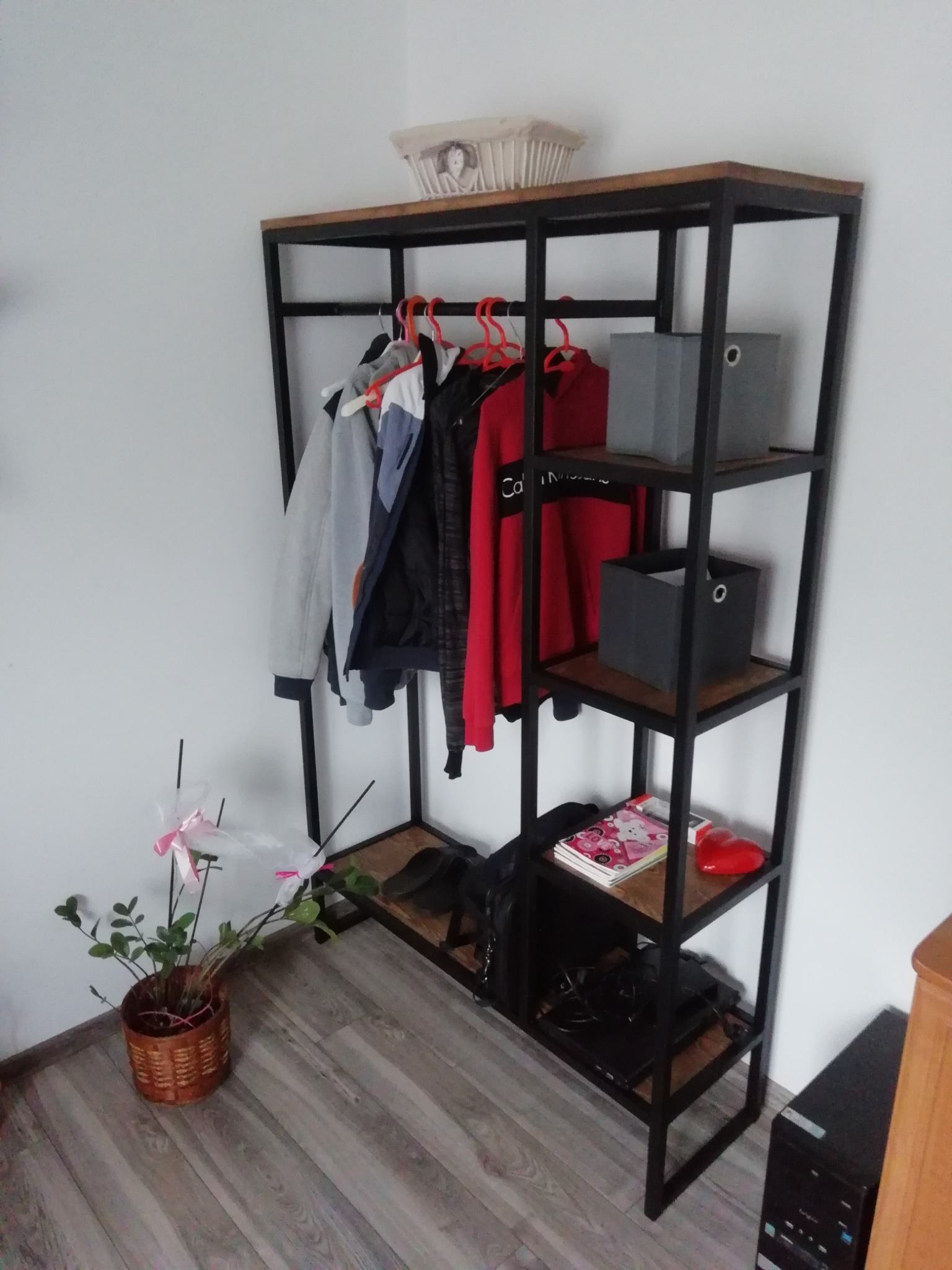 Metalowy regał z drewnianymi półkami, używany jako garderoba, z ubraniami na wieszakach, pudełkami i dekoracjami, ustawiony w rogu pokoju.