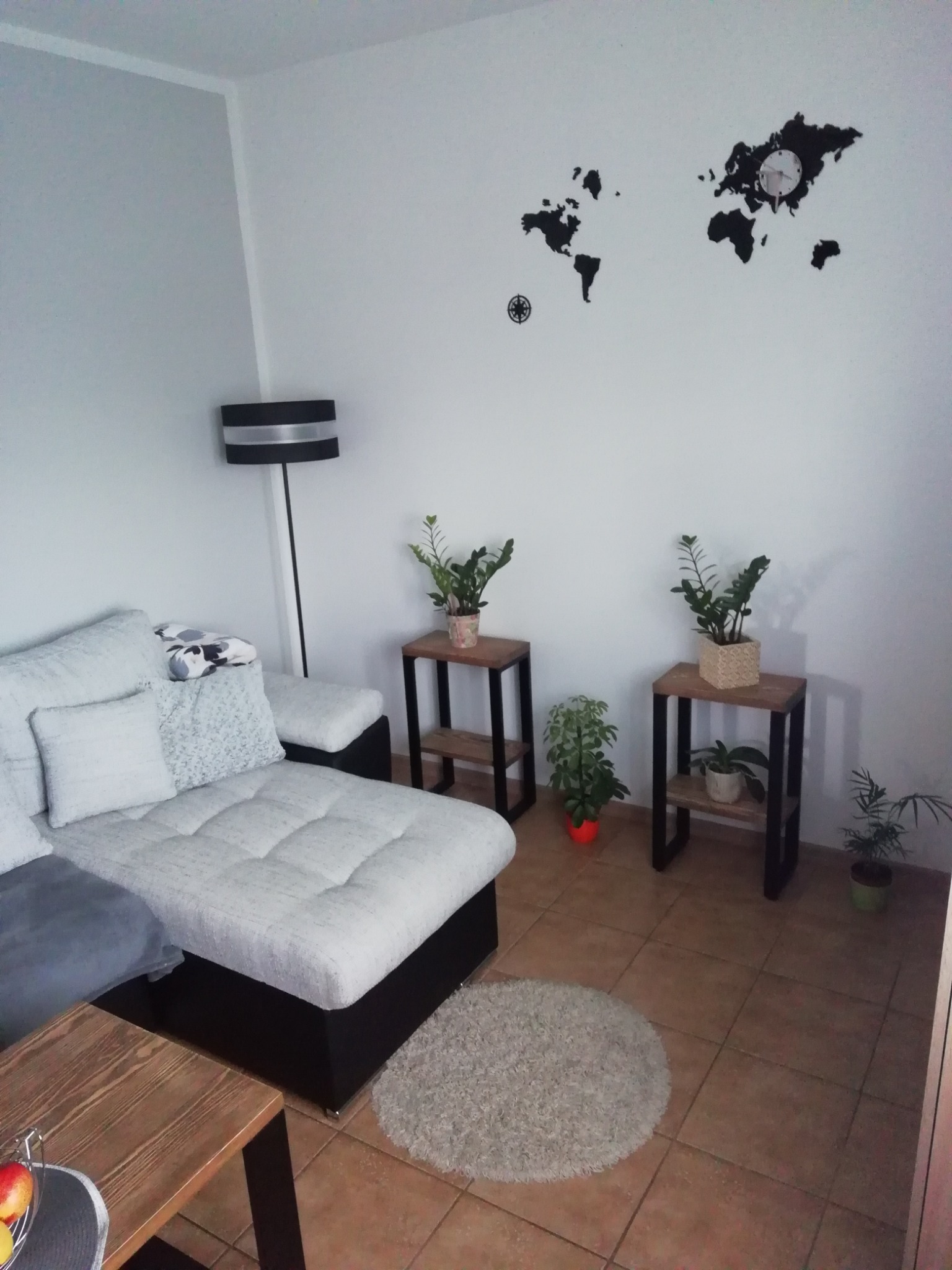 Jasny salon z narożnikiem, lampą podłogową, stolikami z roślinami i dekoracją ścienną w postaci mapy świata z zegarem.