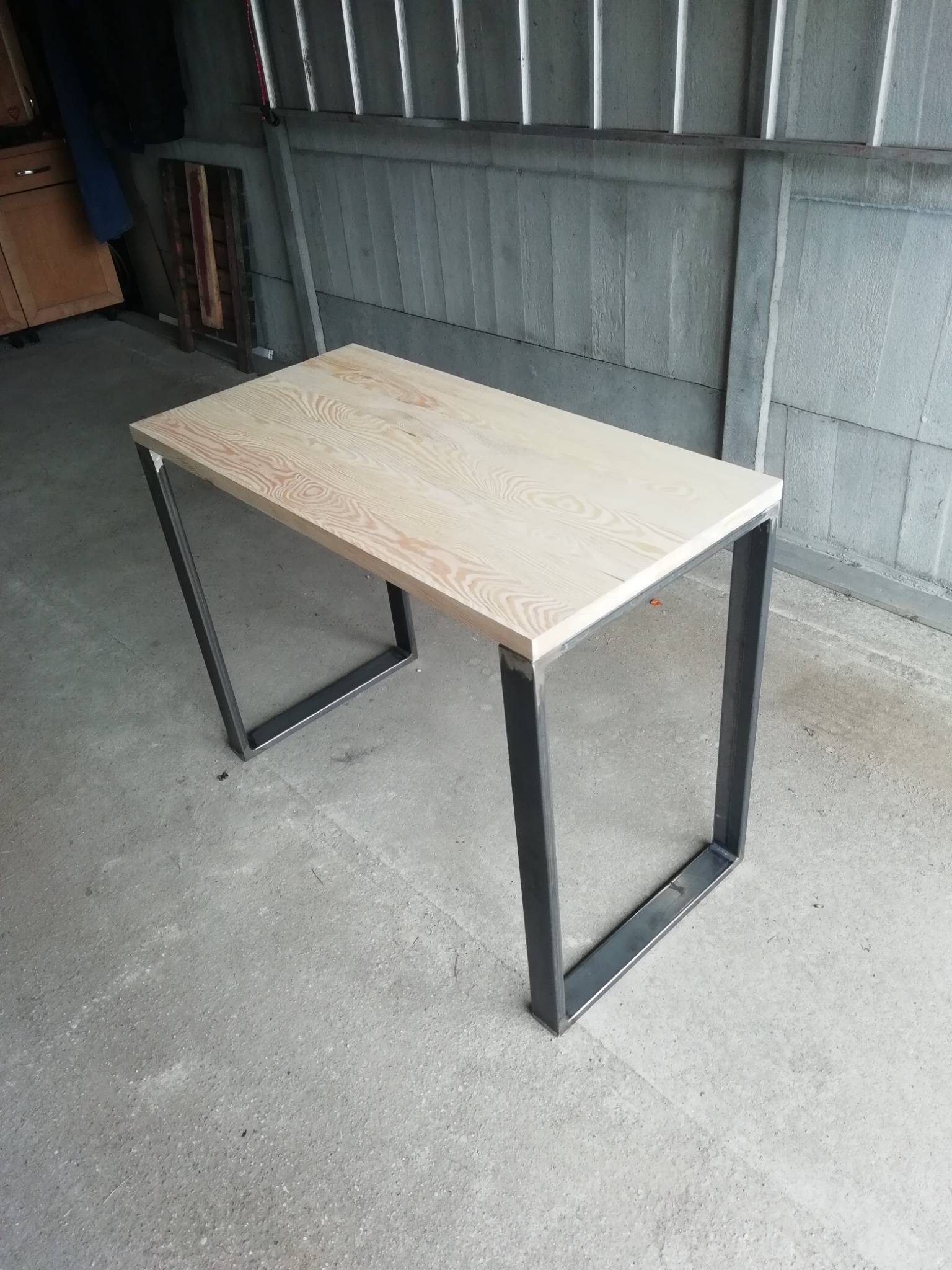 Stół z blatem z jasnego drewna i nogami z profili stalowych o kwadratowym przekroju, spawanych w kształt litery U, surowe otoczenie warsztatowe.