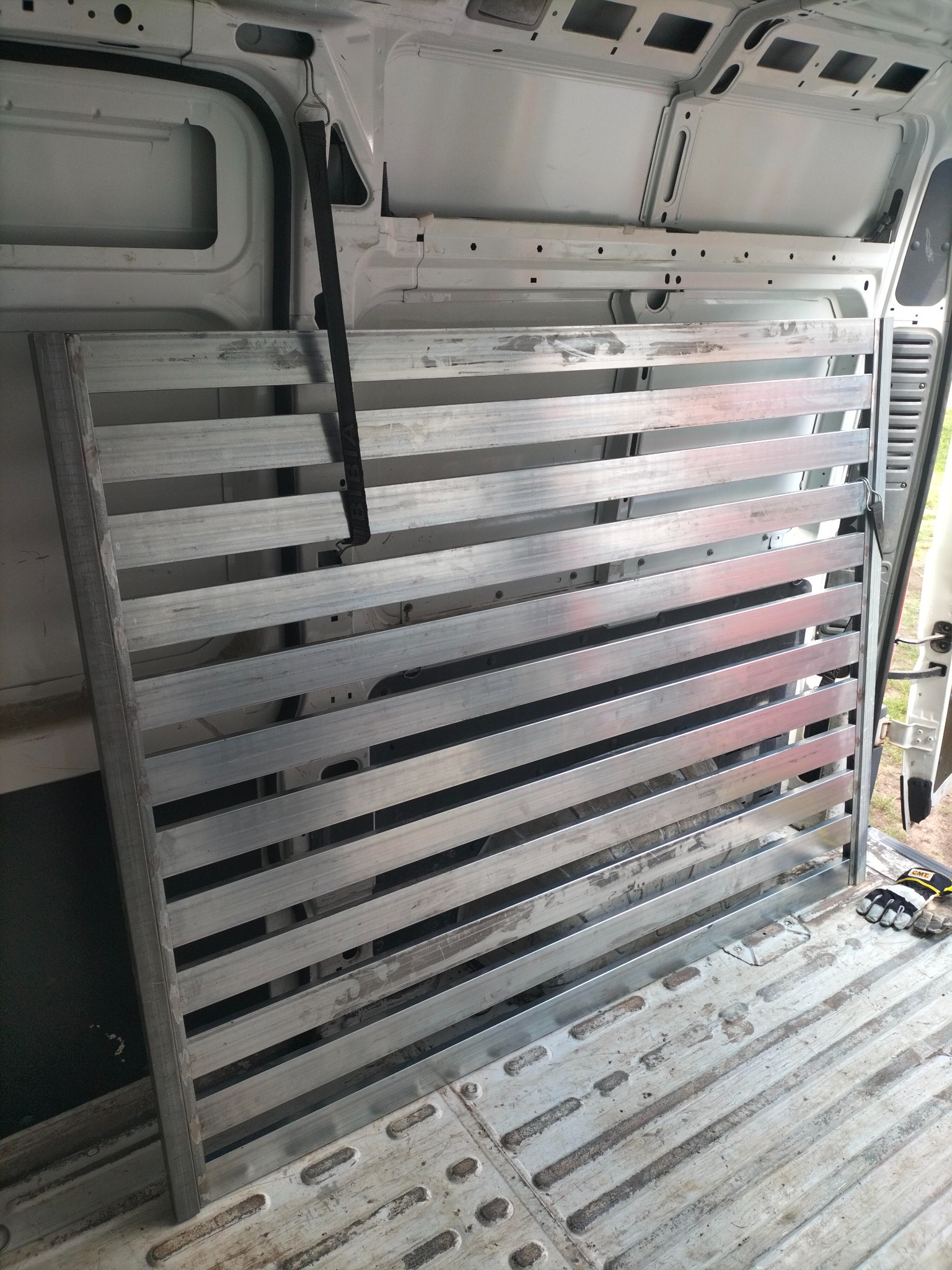 Metalowa kratownica zabezpieczona pasem transportowym wewnątrz białej furgonetki, widoczne rękawice robocze.