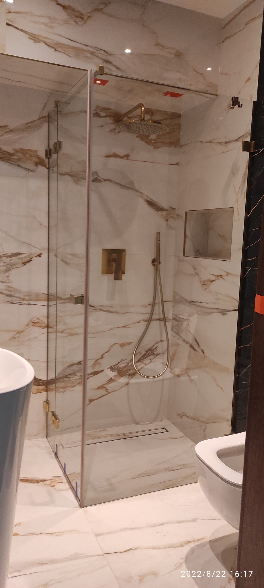 Nowoczesna kabina prysznicowa walk-in z armaturą w kolorze złotym i marmurowymi płytkami na ścianach i podłodze, widoczny odpływ liniowy.