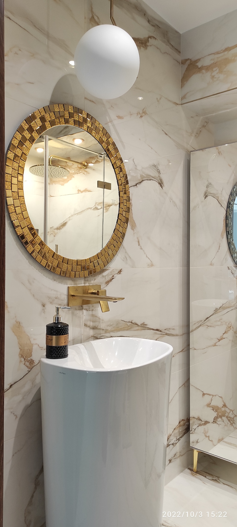 Elegancka łazienka z umywalką na postumencie, złotą armaturą i lustrem w mozaikowej ramie na tle marmurowych płytek. W lustrze odbija się kabina prysznicowa.
