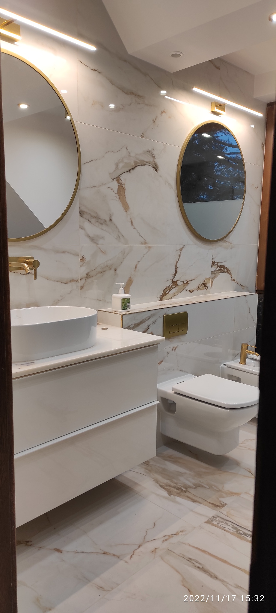 Nowoczesna łazienka z białymi płytkami imitującymi marmur, okrągłymi lustrami w złotych ramach, białą umywalką nablatową, toaletą i bidetem, z minimalistyczną armaturą w kolorze złotym.