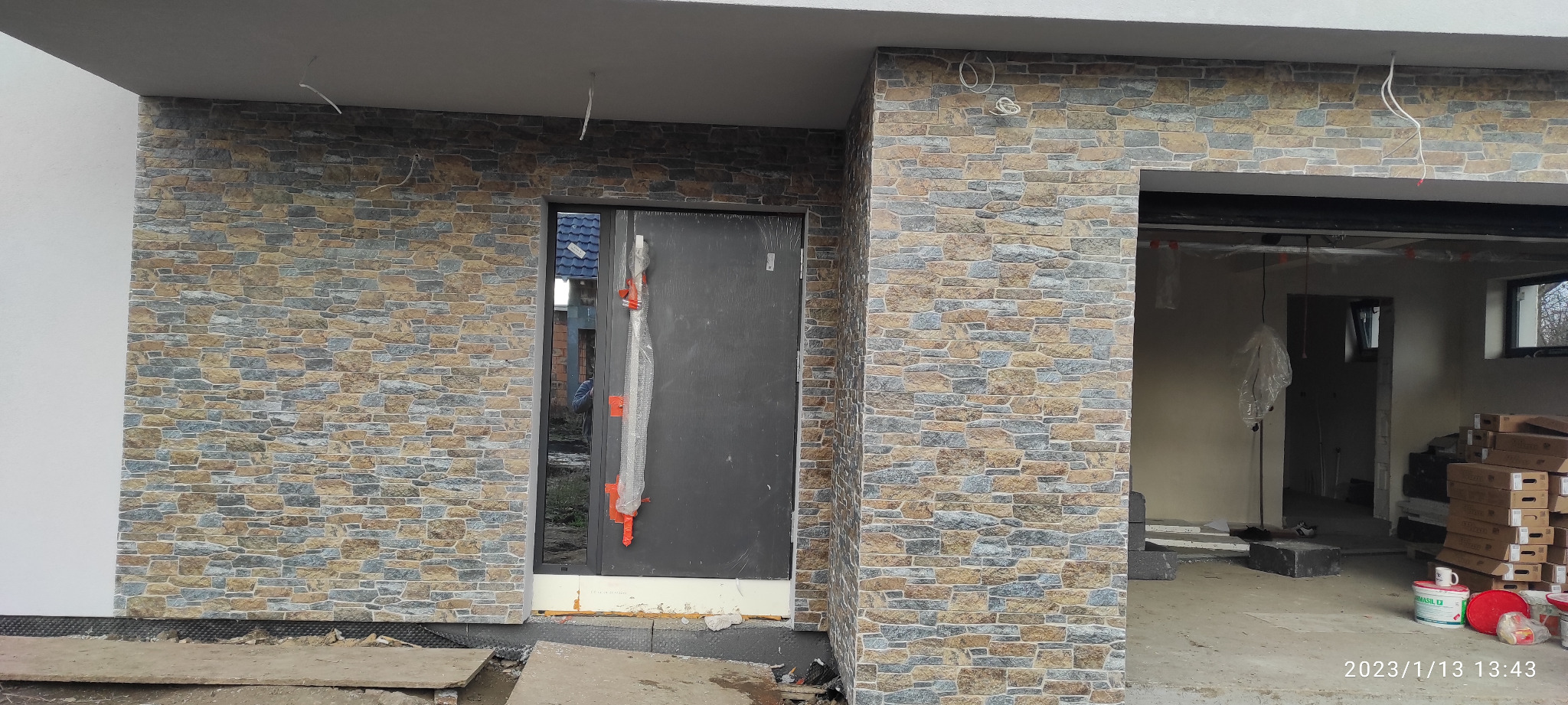 Elewacja budynku wykończona płytkami imitującymi kamień, widoczne drzwi wejściowe z zabezpieczeniem folią i niezakończone prace wykończeniowe wewnątrz budynku.