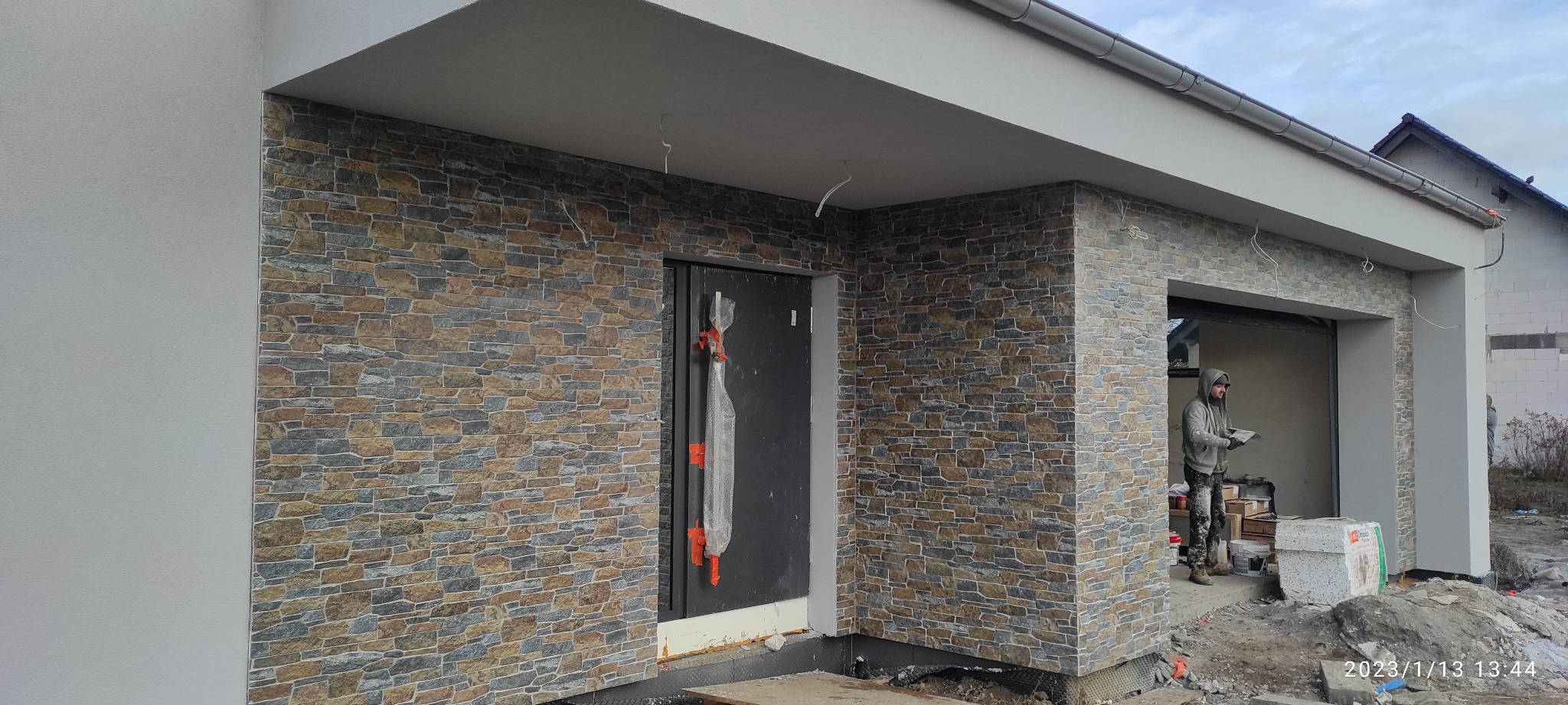 Elewacja budynku z kamienia dekoracyjnego w odcieniach brązu i szarości, widoczny pracownik w trakcie wykonywania prac wykończeniowych, zabezpieczone okno folią ochronną.