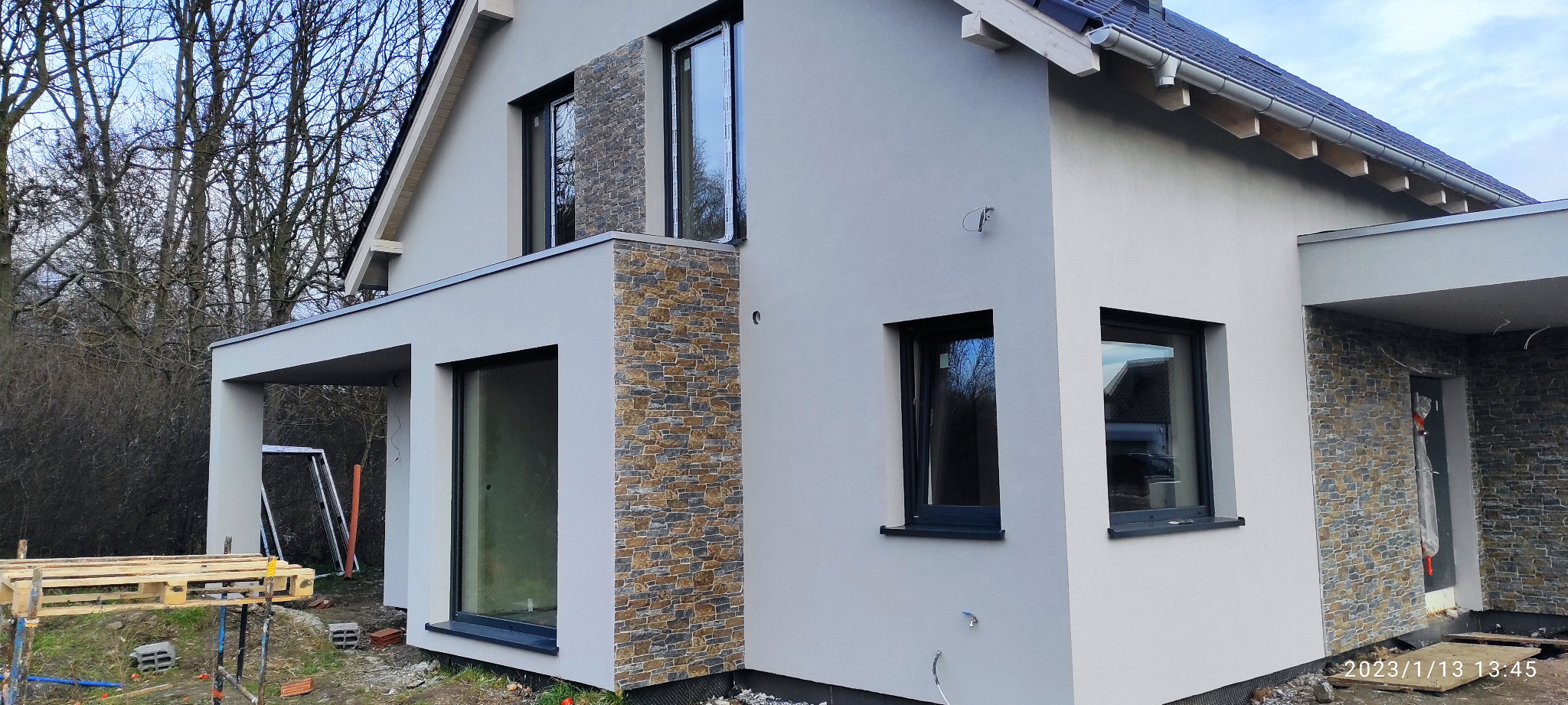 Nowoczesny dom jednorodzinny z fragmentami elewacji wykończonymi kamieniem dekoracyjnym, widoczne okna z czarnymi ramami, paleta z materiałami budowlanymi przed budynkiem.