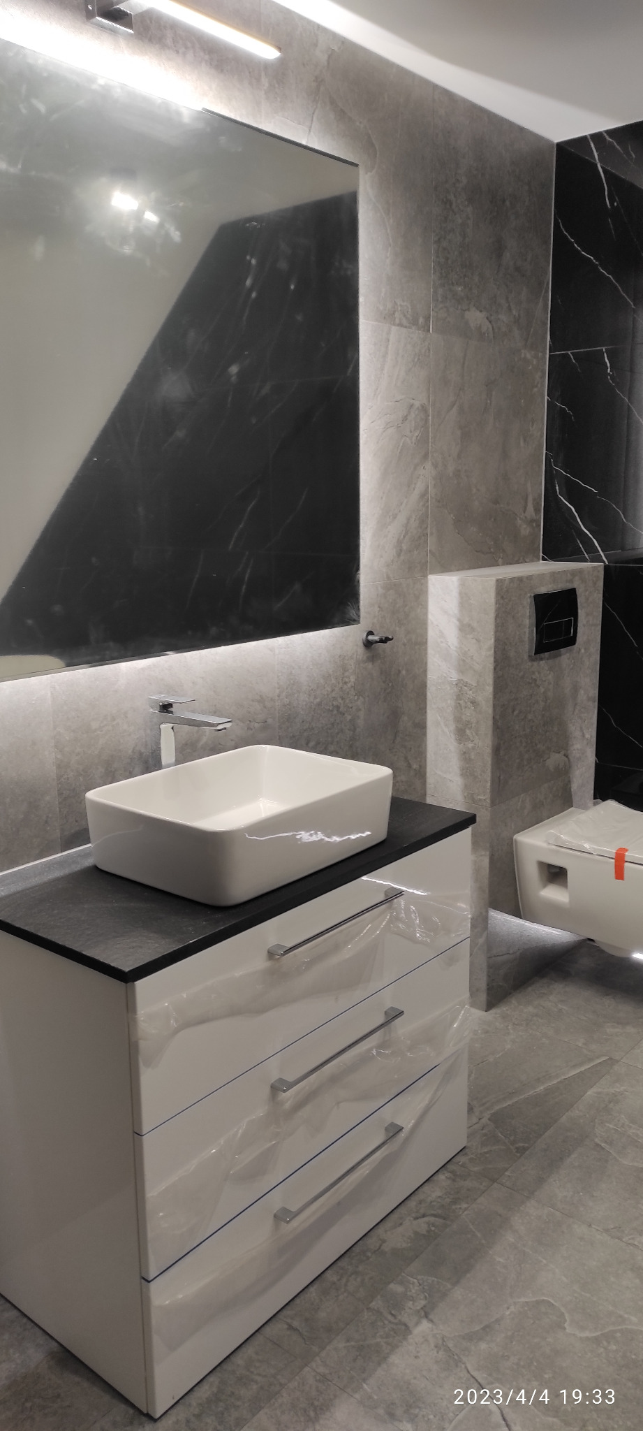 Nowoczesna łazienka z umywalką nablatową na białej szafce z trzema szufladami, szare płytki imitujące marmur na ścianach oraz wisząca miska WC z ukrytym systemem spłukiwania, całość jeszcze...