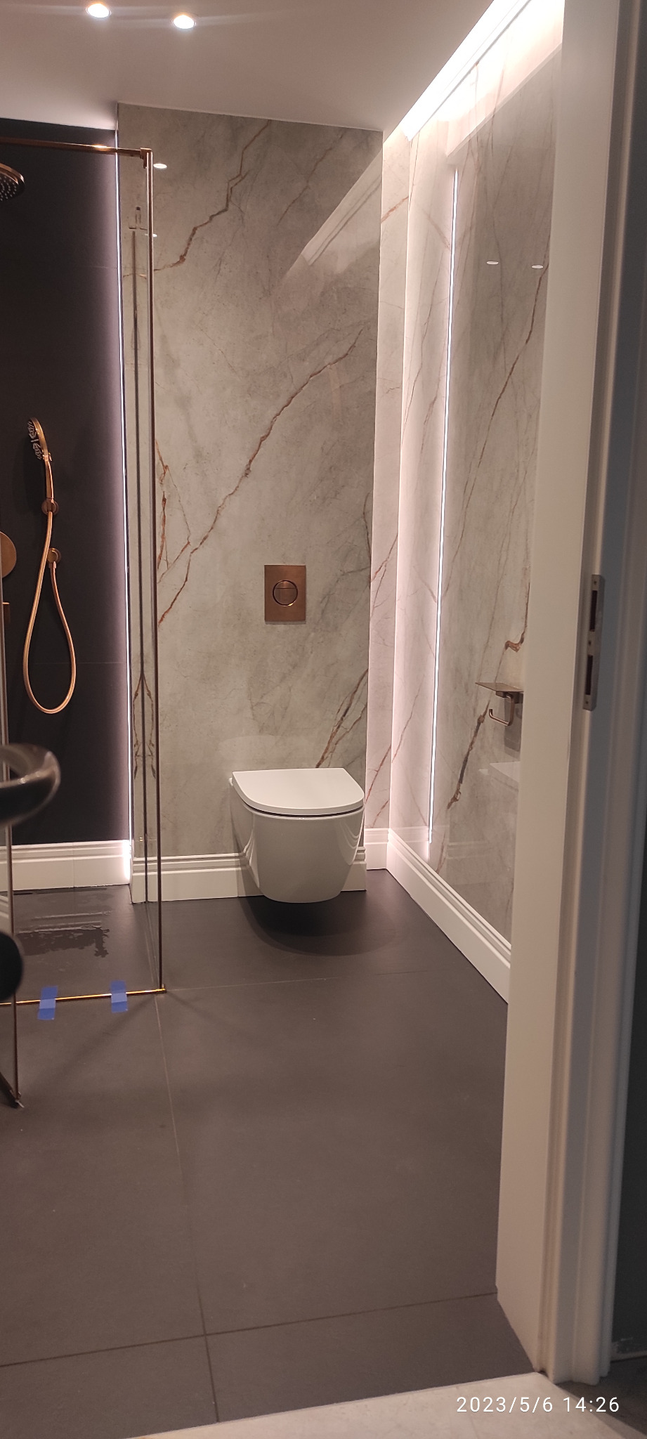 Nowoczesna łazienka z marmurowymi płytkami ściennymi, podwieszaną toaletą i kabiną prysznicową z elementami w kolorze miedzi.