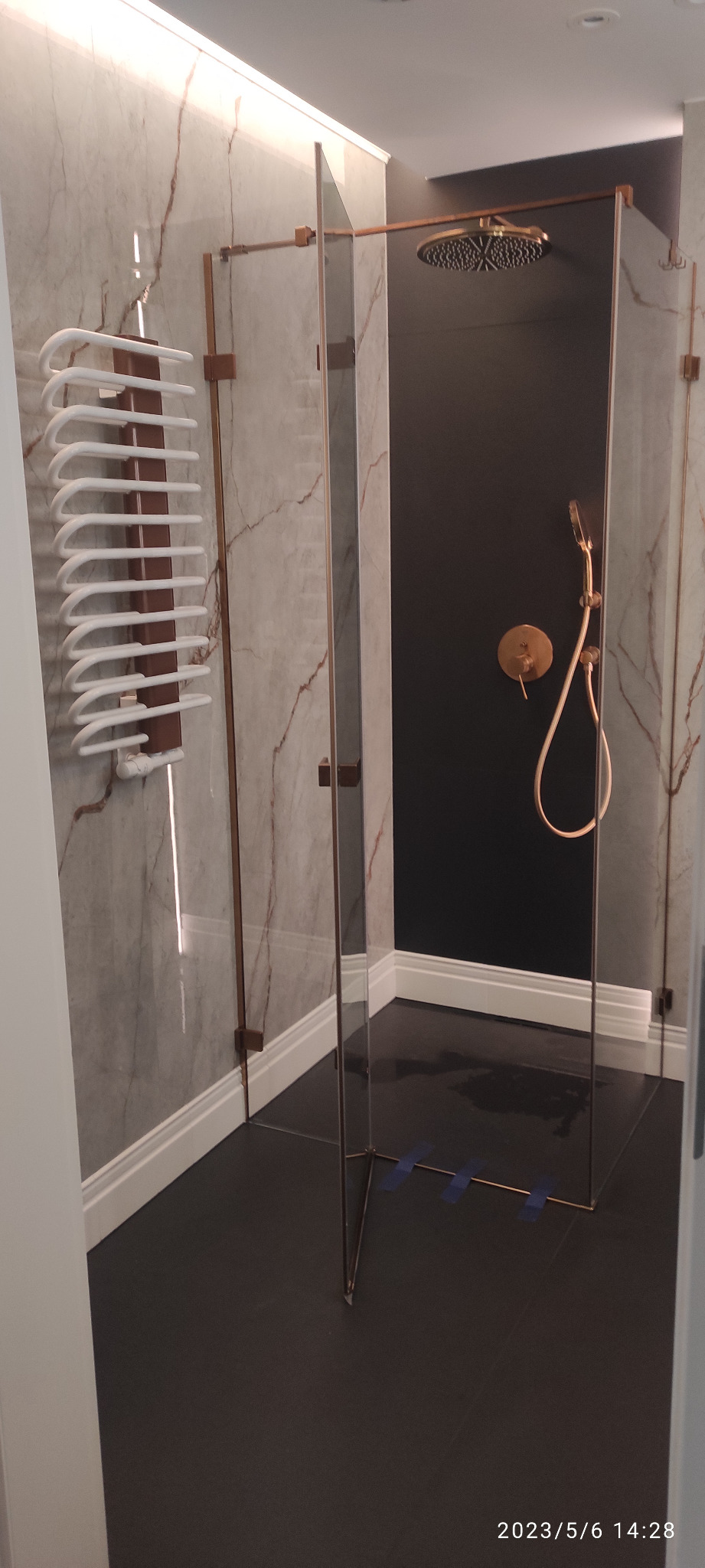 Nowoczesna kabina prysznicowa z marmurowymi ścianami, deszczownicą i grzejnikiem drabinkowym, widoczne taśmy zabezpieczające na podłodze.