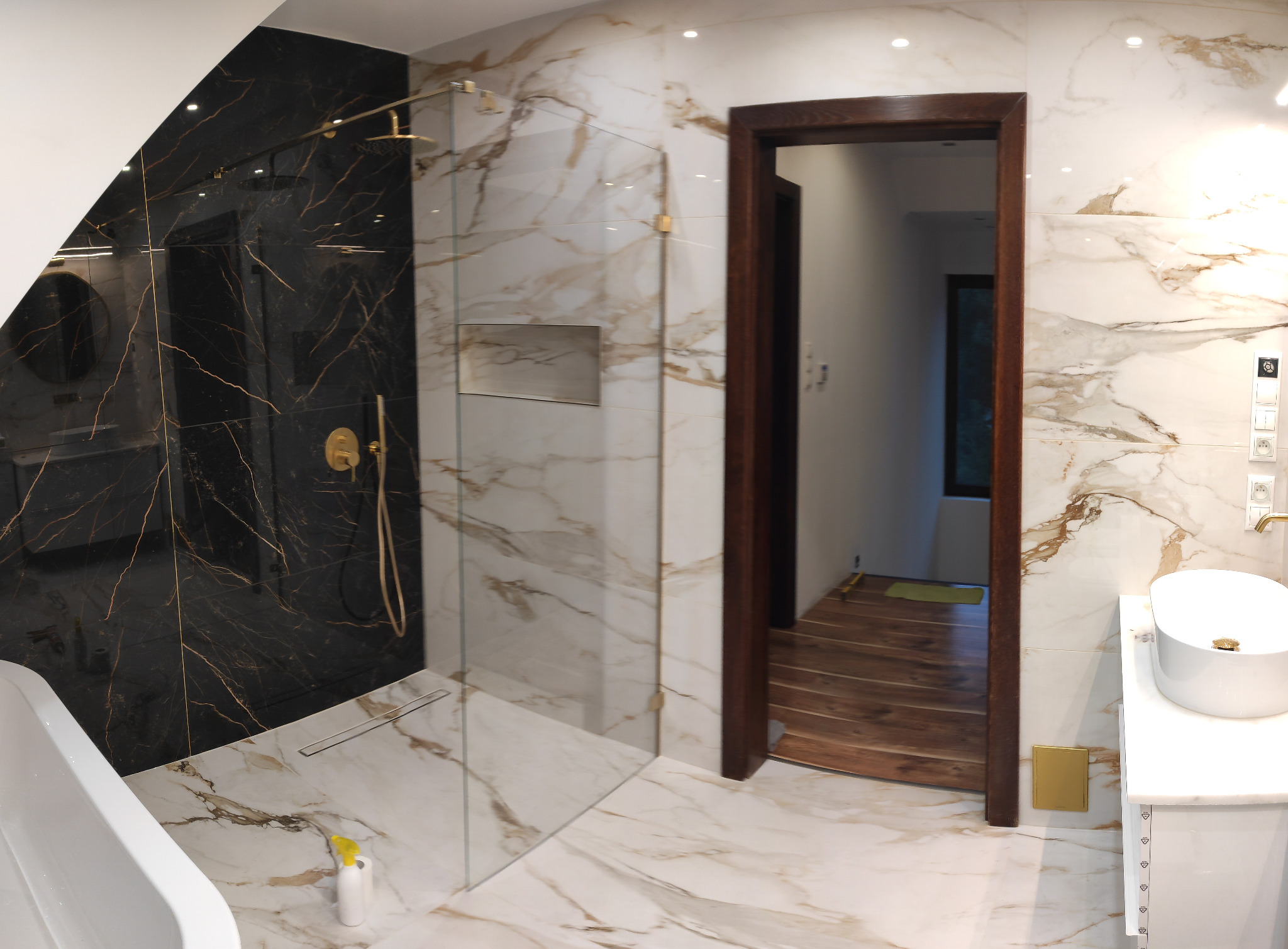 Elegancka łazienka z kabiną prysznicową typu walk-in wyłożoną czarnymi płytkami z złotymi żyłami, marmurowe płytki na ścianach i podłodze, biała umywalka nablatowa i widok na drewnianą podłogę...