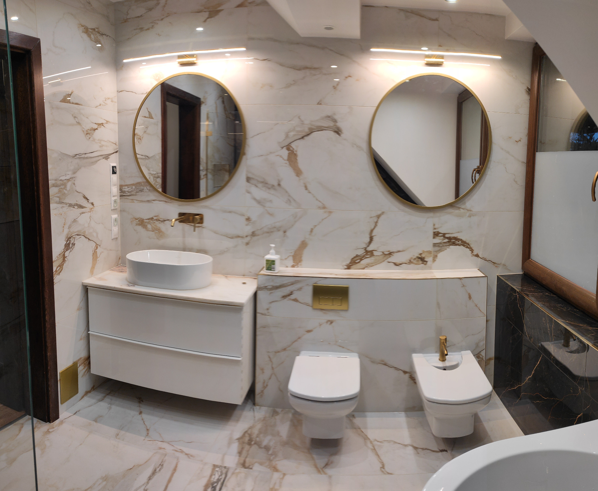 Elegancka łazienka z marmurowymi płytkami, okrągłymi lustrami w złotych ramach, białą umywalką nablatową i wiszącą toaletą oraz bidetem z baterią w kolorze złotym. Widoczne elementy wykończeniowe...