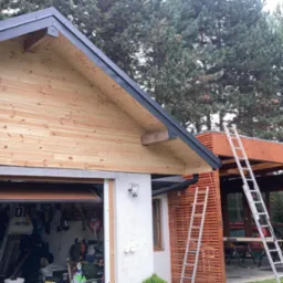 Wykończona drewnianą boazerią ściana zewnętrzna garażu z szarą metalową obróbką dachu, z otwartą bramą garażową i widocznymi narzędziami w środku, obok dobudowana drewniana altana z drabina opartą...