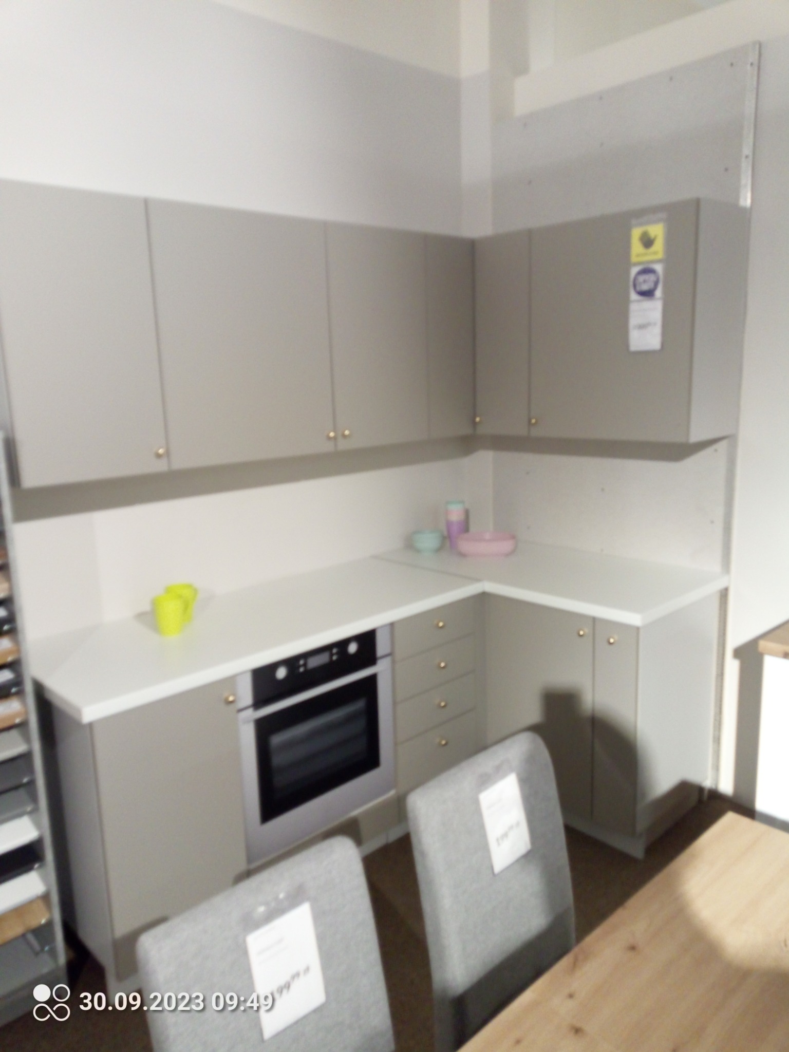 Jasnoszara zabudowa kuchenna z wbudowanym piekarnikiem, z blatem w kolorze bieli i złotymi uchwytami, widok z perspektywy krzesła z ceną.