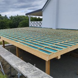 Tartacznictwo Usługi-Produkcja - Konstrukcja drewniana z widoczną izolacją w kolorze niebieskim, prawdopodobnie pod przyszły taras, z rusztowaniem obok, na tle białego domu i pochmurnego nieba.