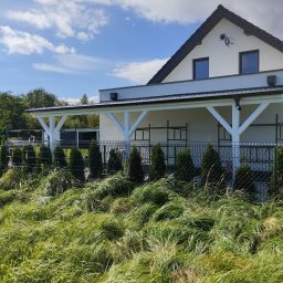 Tartacznictwo Usługi-Produkcja - Biała konstrukcja zadaszenia tarasu przylegająca do domu, widoczna przez ogrodzenie i wysokie trawy, w tle niebo z chmurami.