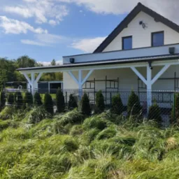 Biała konstrukcja zadaszenia tarasu przylegająca do domu, widoczna przez ogrodzenie i wysokie trawy, w tle niebo z chmurami.