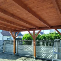 Drewniana konstrukcja z belek i słupów, prawdopodobnie zadaszenie tarasu w trakcie budowy, widok od dołu, z widocznym ogrodzeniem z betonu i kamienia oraz zielenią w tle.