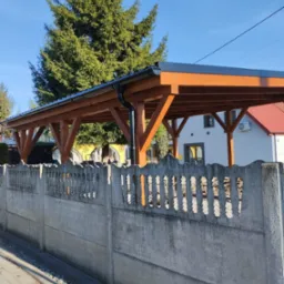 Drewniana pergola z czarnym dachem, widoczna zza betonowego ogrodzenia, z zielonym drzewem w tle.