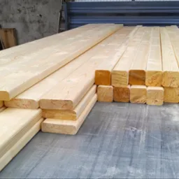 Świeże, jasne drewno konstrukcyjne ułożone w stosy na szarym blacie roboczym. Widoczne ślady obróbki drewna i niewielkie czerwone oznaczenia na kantach.