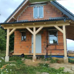 Drewniana konstrukcja zadaszenia przy murowanym domu w trakcie budowy, widoczne elementy więźby dachowej i fragment krajobrazu w tle.