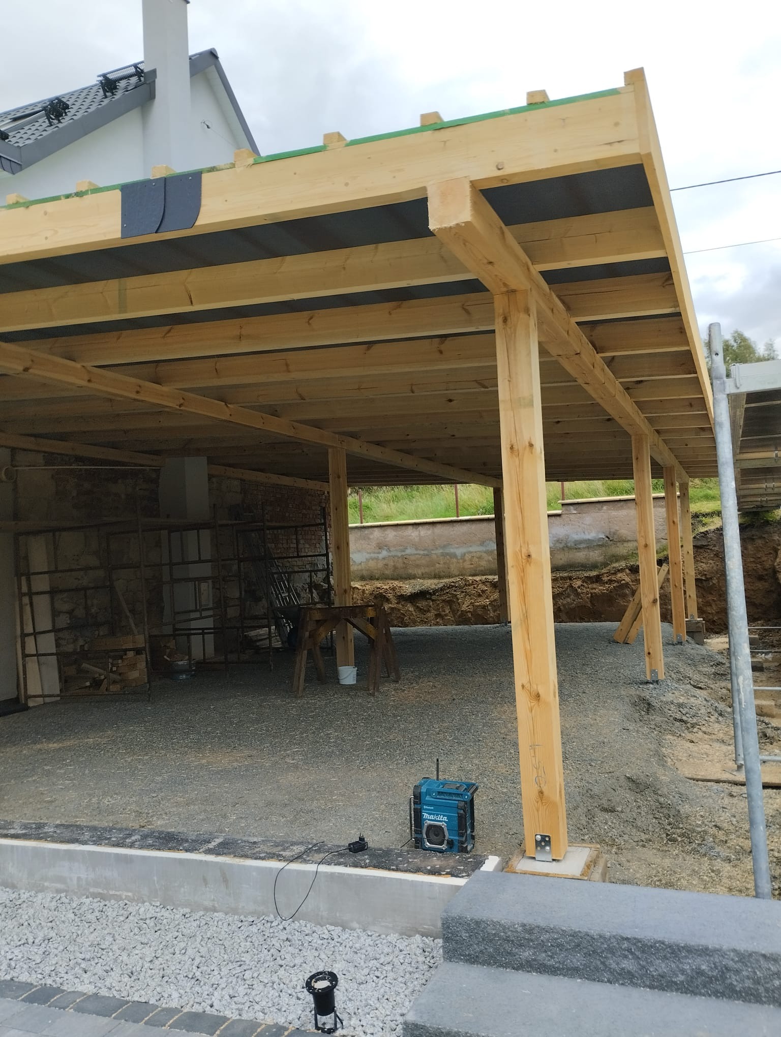 Konstrukcja z surowego drewna, prawdopodobnie zadaszenie tarasu lub wiaty, w trakcie budowy; widoczny betonowy fundament, podłoże z kamieni, narzędzia i radio budowlane.