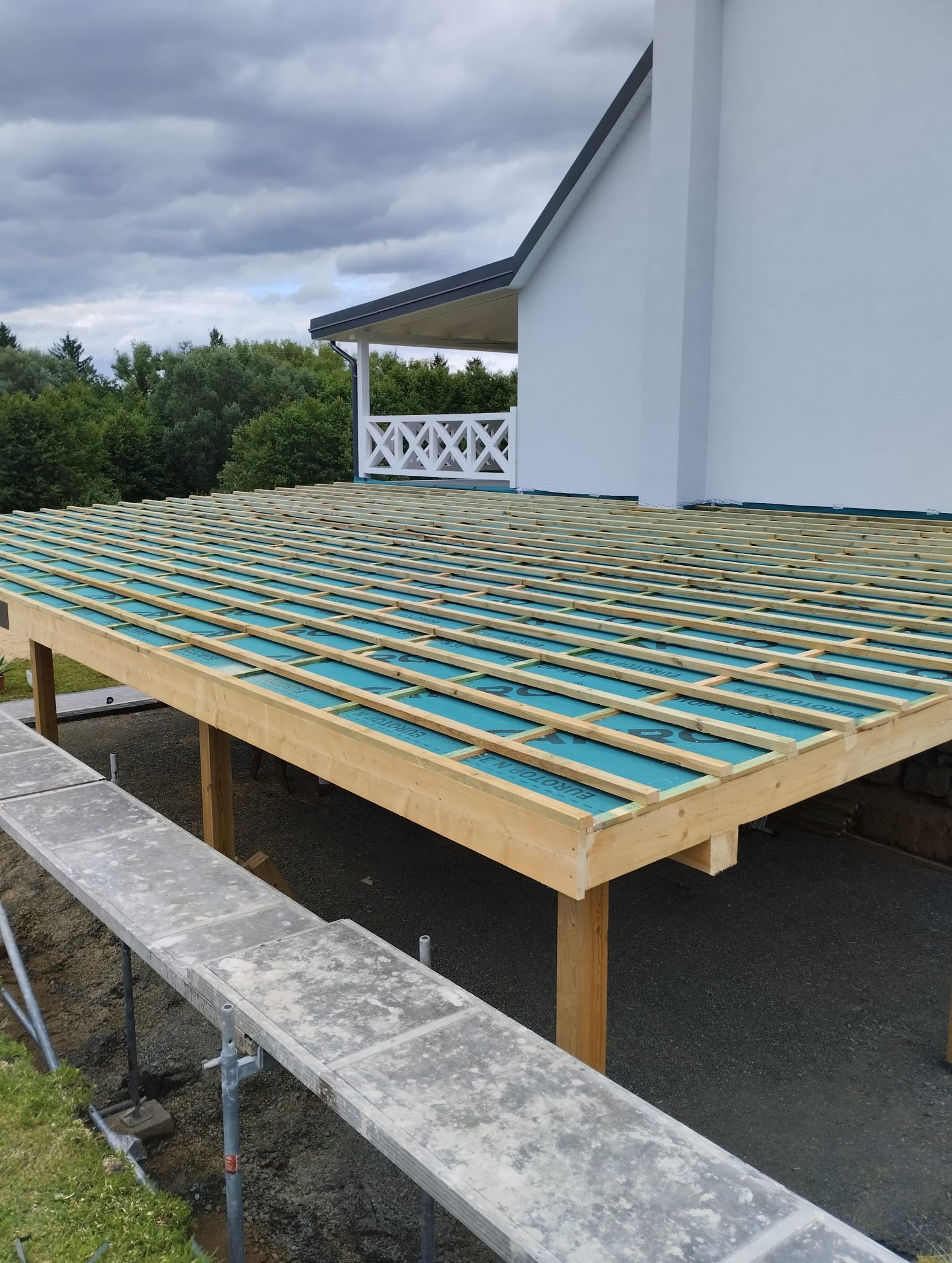 Konstrukcja drewniana z widoczną izolacją w kolorze niebieskim, prawdopodobnie pod przyszły taras, z rusztowaniem obok, na tle białego domu i pochmurnego nieba.
