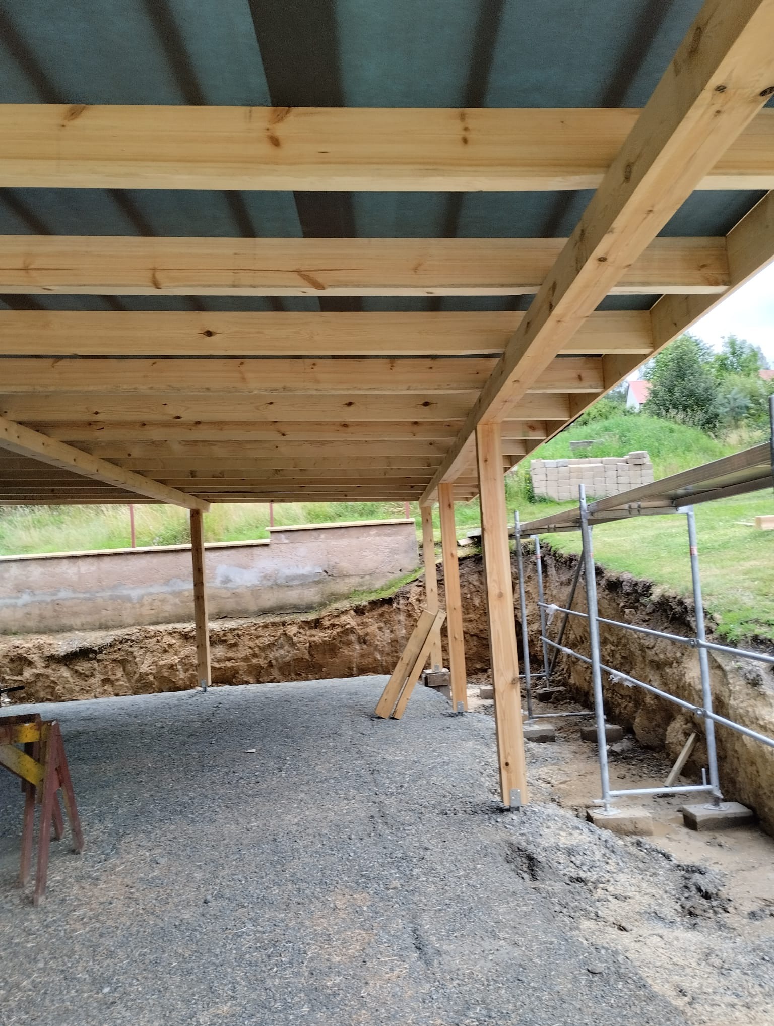 Konstrukcja drewniana z widocznymi belkami i słupami, prawdopodobnie w trakcie budowy tarasu lub zadaszenia, z widocznym wykopem i rusztowaniem w tle.