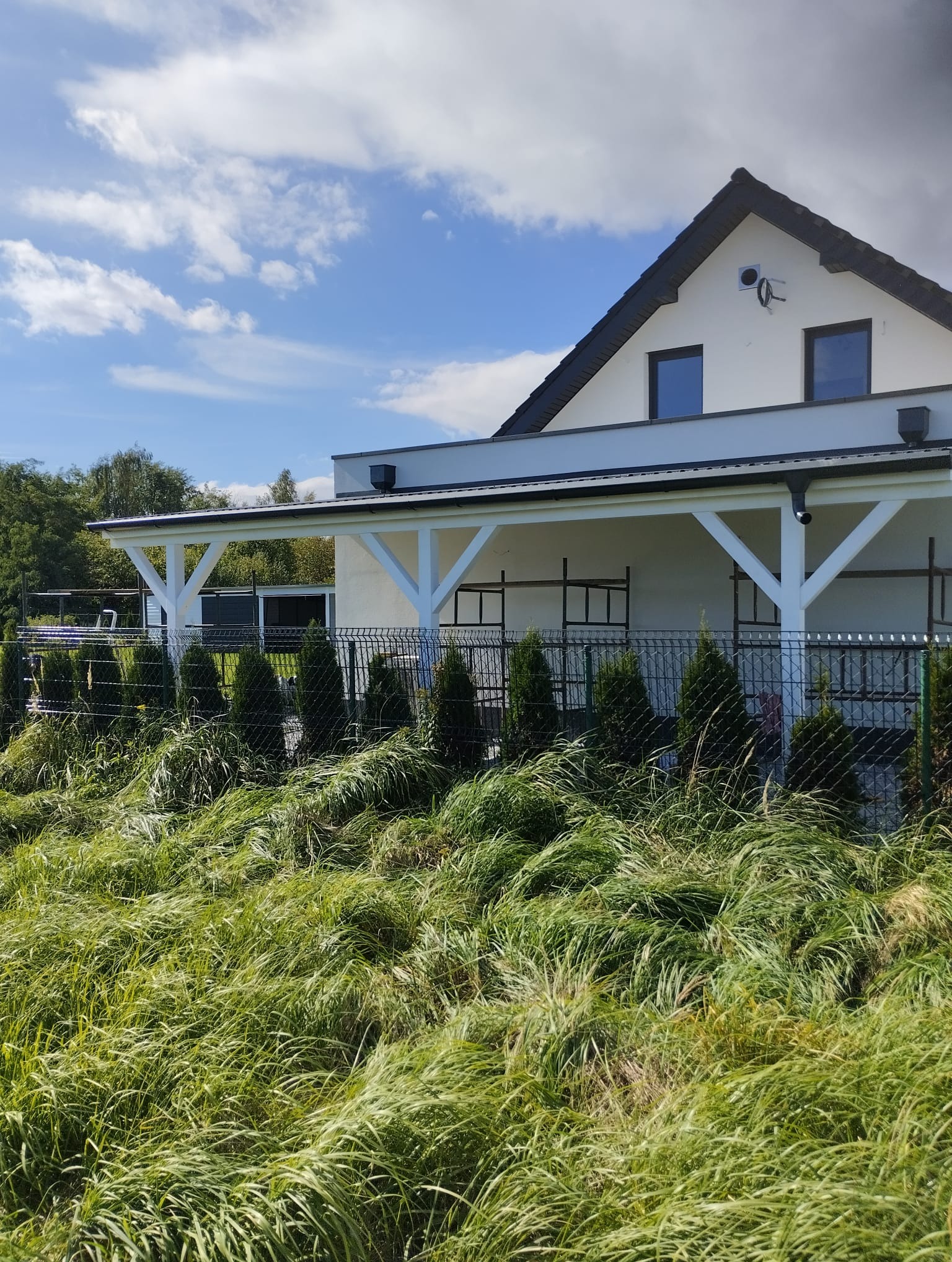 Biała konstrukcja zadaszenia tarasu przylegająca do domu, widoczna przez ogrodzenie i wysokie trawy, w tle niebo z chmurami.