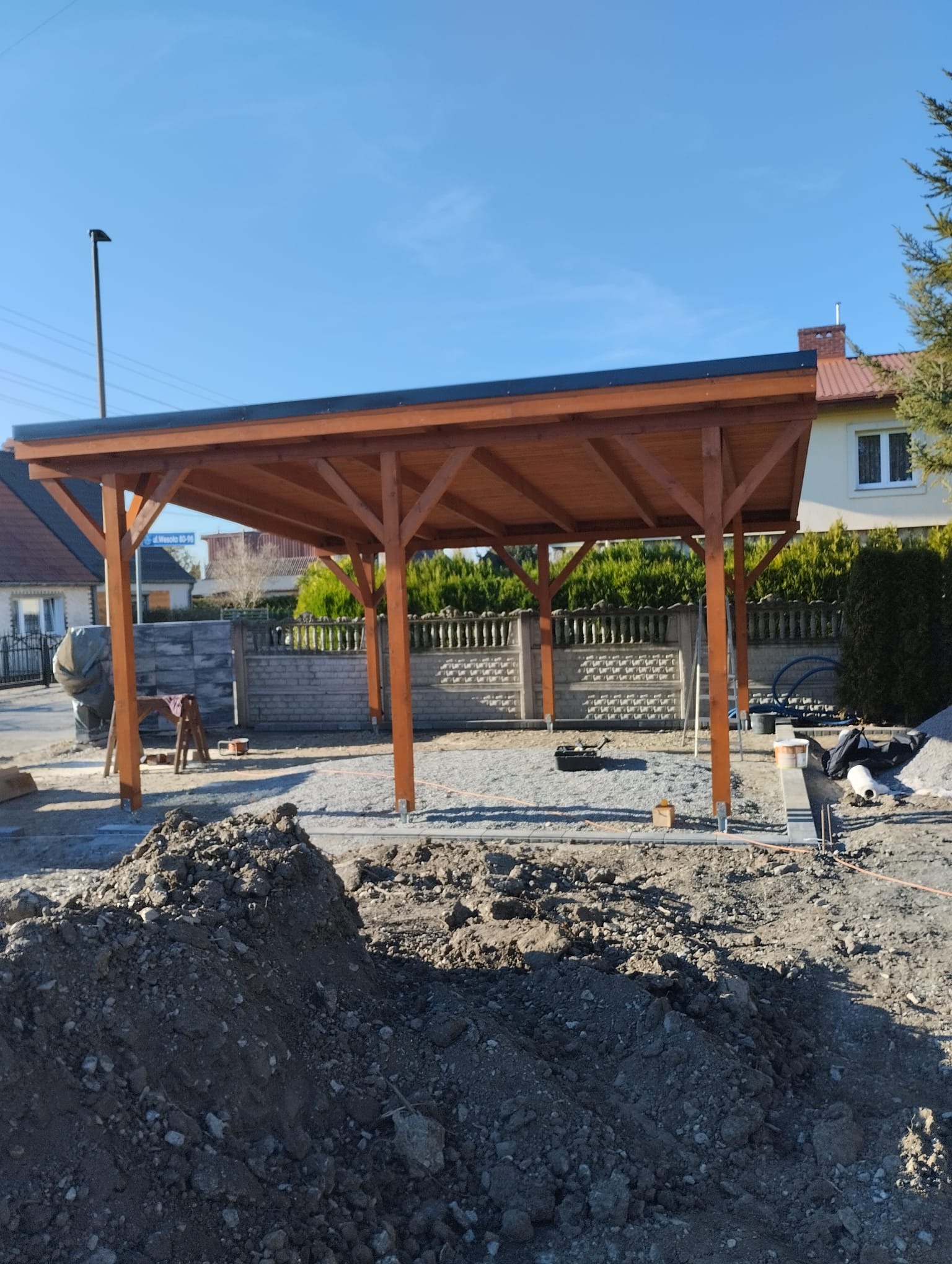 Drewniana konstrukcja z czarnym dachem w trakcie montażu, widoczne fundamenty i przygotowany teren.