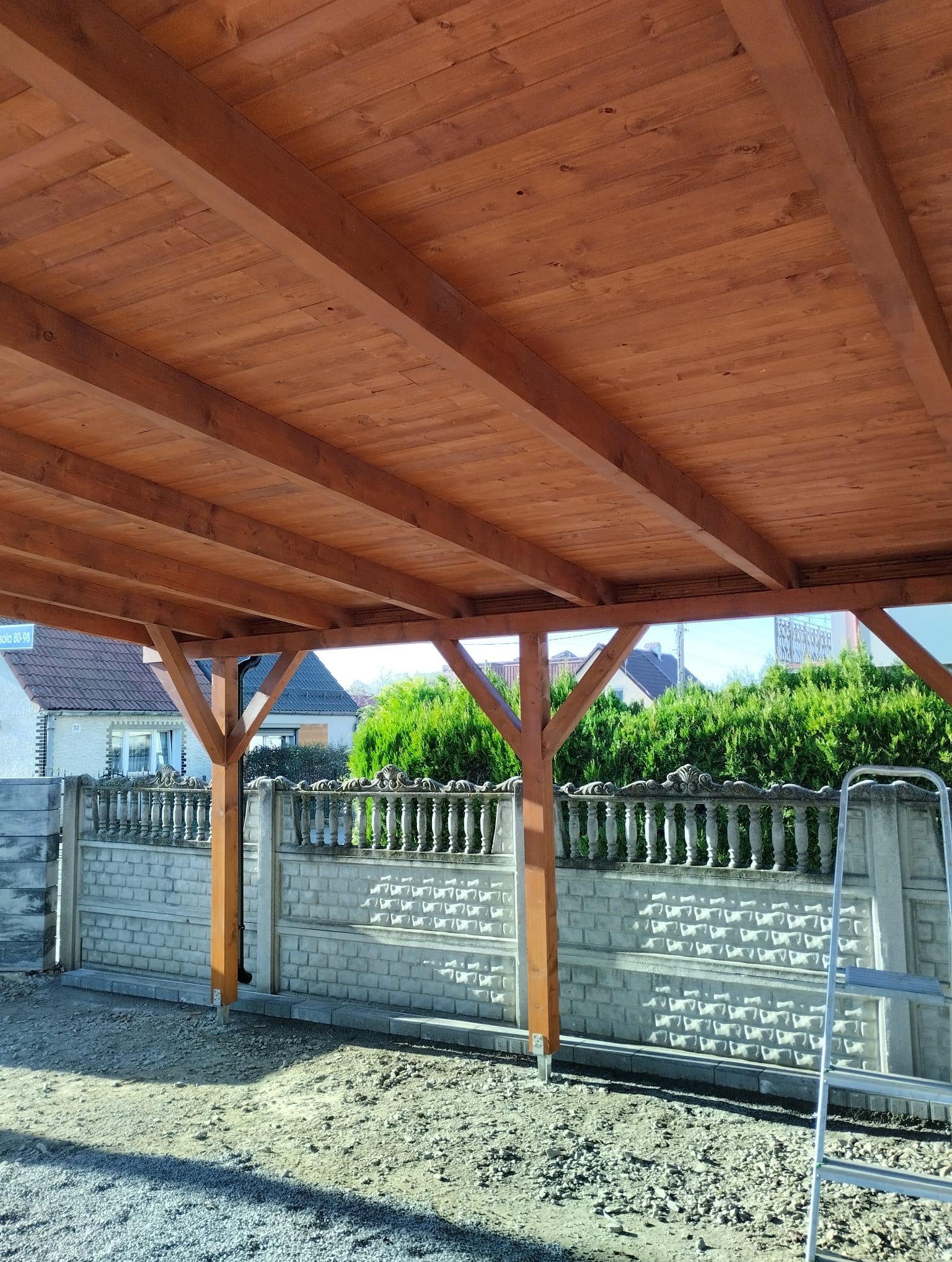 Drewniana konstrukcja z belek i słupów, prawdopodobnie zadaszenie tarasu w trakcie budowy, widok od dołu, z widocznym ogrodzeniem z betonu i kamienia oraz zielenią w tle.