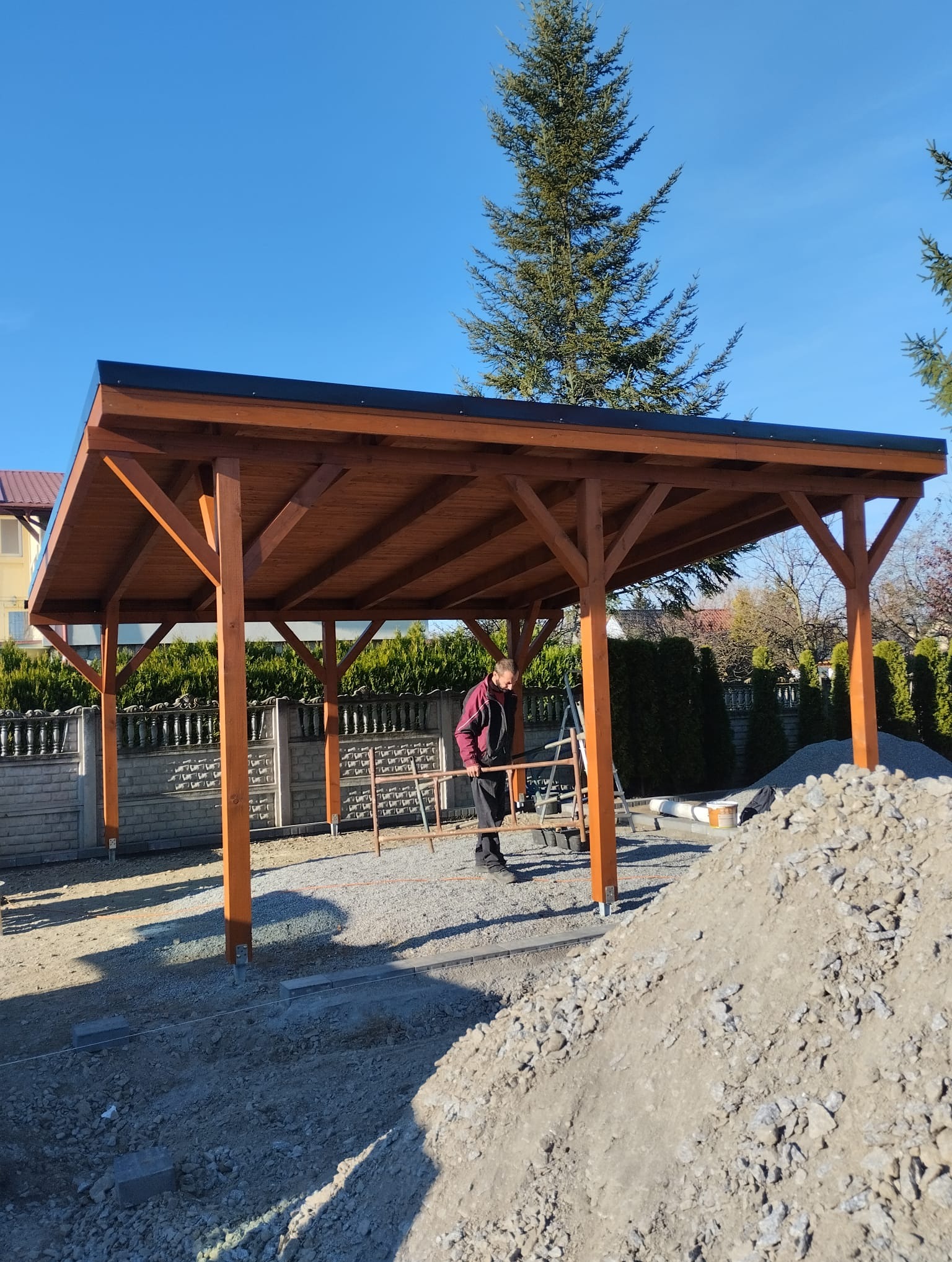 Montaż drewnianej konstrukcji z czarnym dachem na przygotowanym terenie, widoczny mężczyzna przy pracy z poziomicą, w tle ogrodzenie z betonowych elementów i iglaste drzewa.