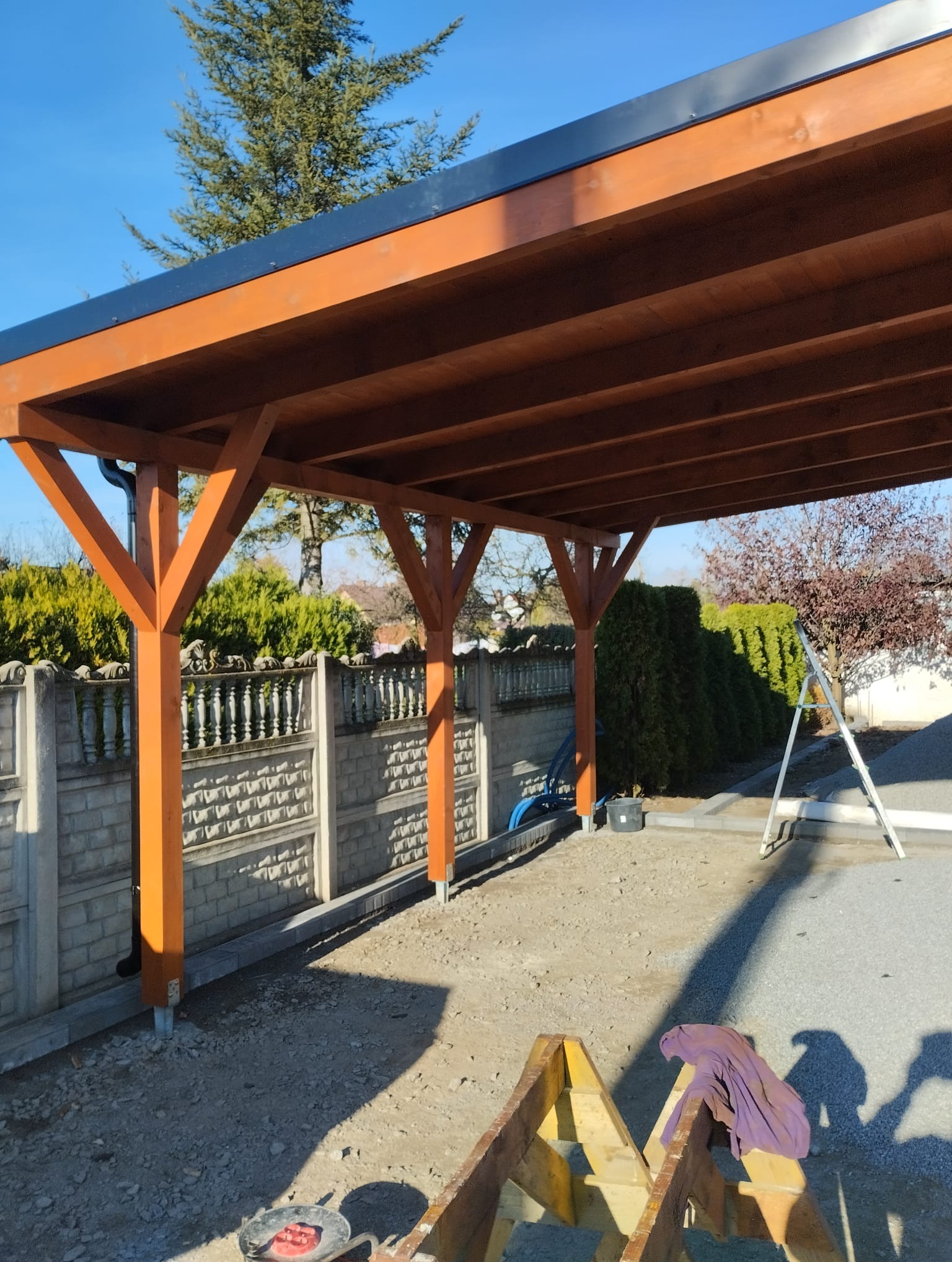 Konstrukcja z belek drewnianych z czarnym dachem, osadzona na metalowych kotwach w gruncie, widoczna na tle ogrodzenia z betonu i zielonych krzewów.