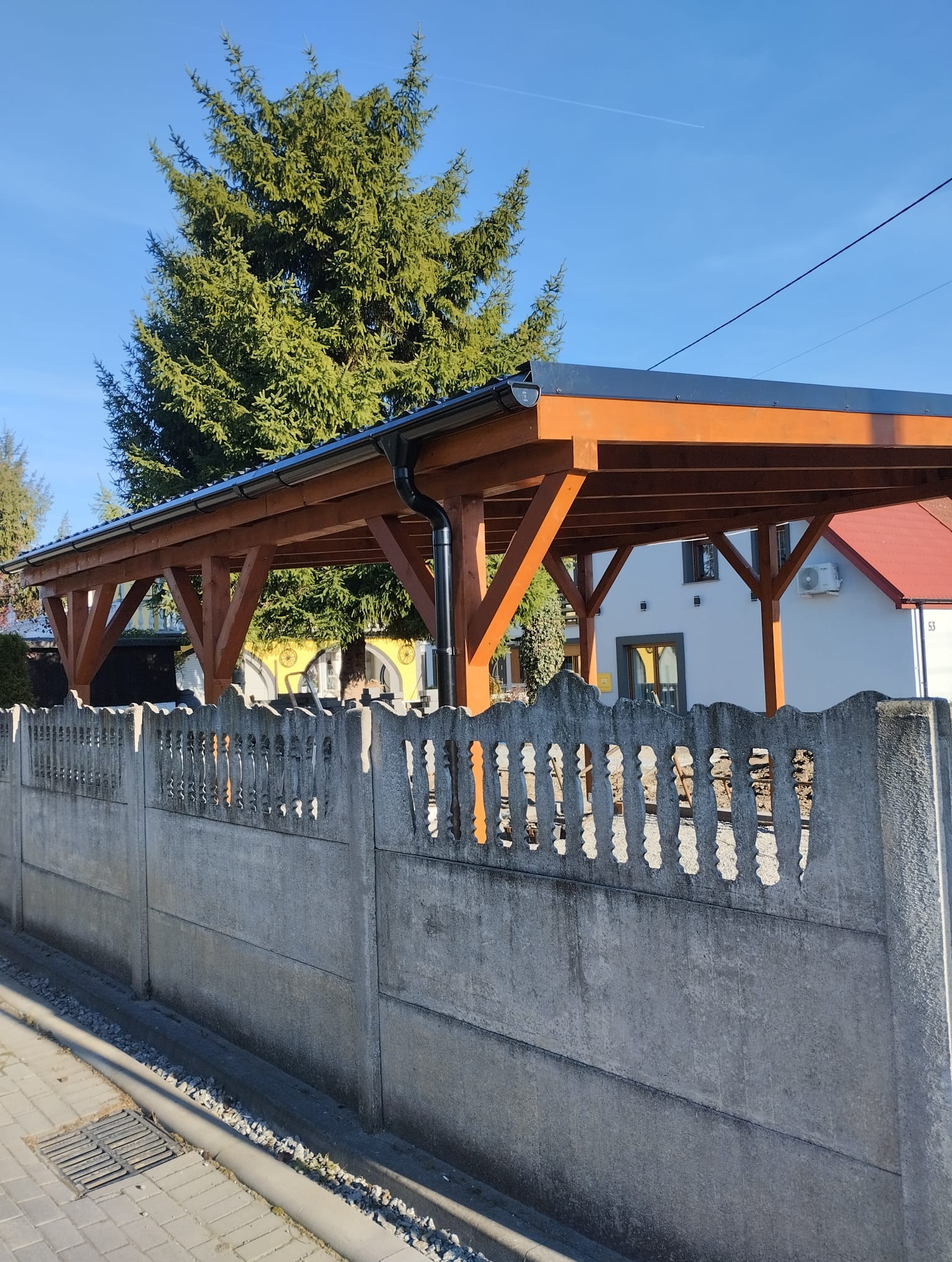 Drewniana pergola z czarnym dachem, widoczna zza betonowego ogrodzenia, z zielonym drzewem w tle.