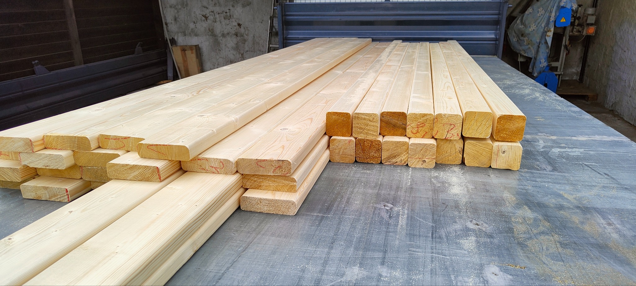 Świeże, jasne drewno konstrukcyjne ułożone w stosy na szarym blacie roboczym. Widoczne ślady obróbki drewna i niewielkie czerwone oznaczenia na kantach.