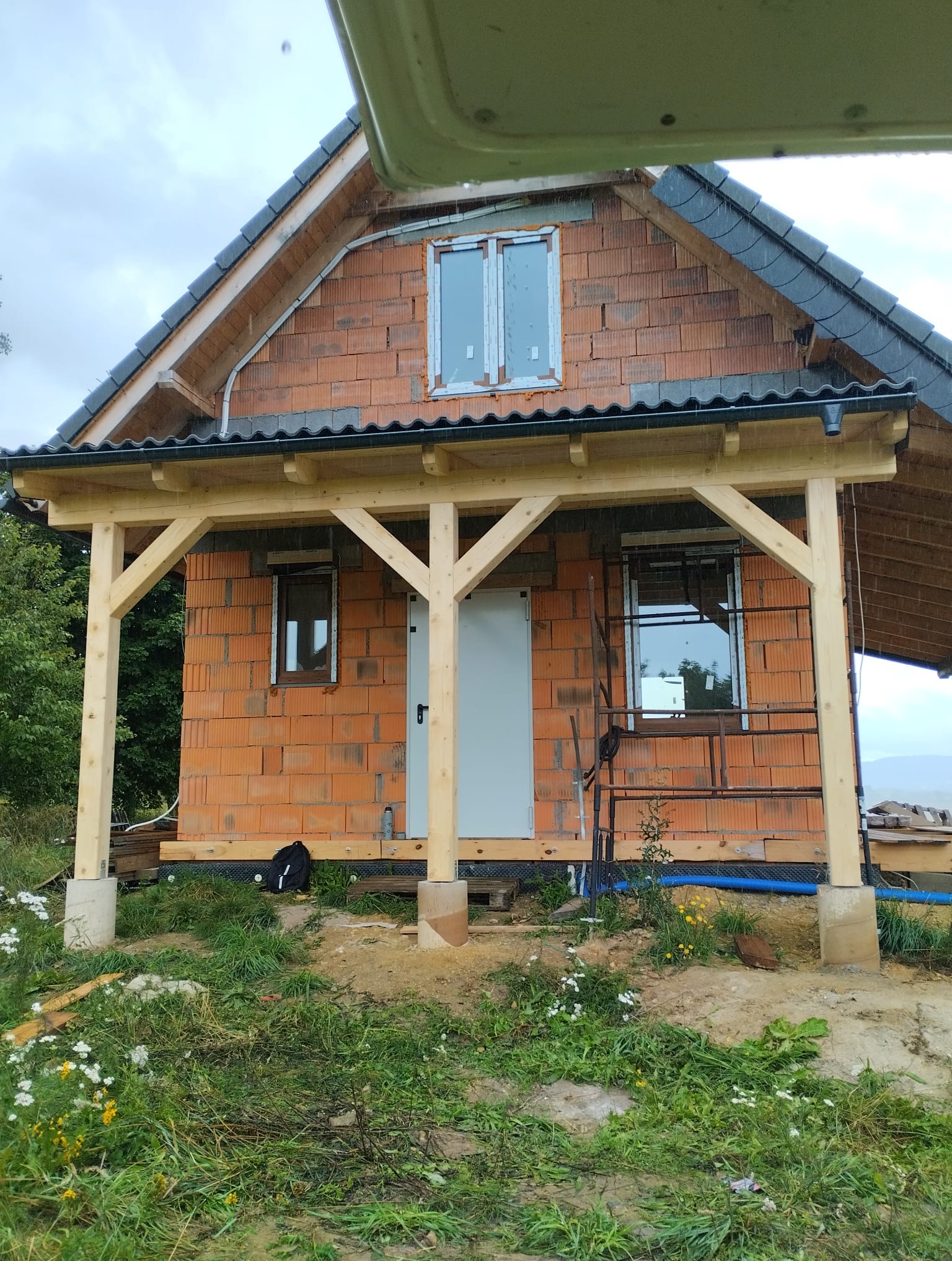 Drewniana konstrukcja zadaszenia przy murowanym domu w trakcie budowy, widoczne elementy więźby dachowej i fragment krajobrazu w tle.