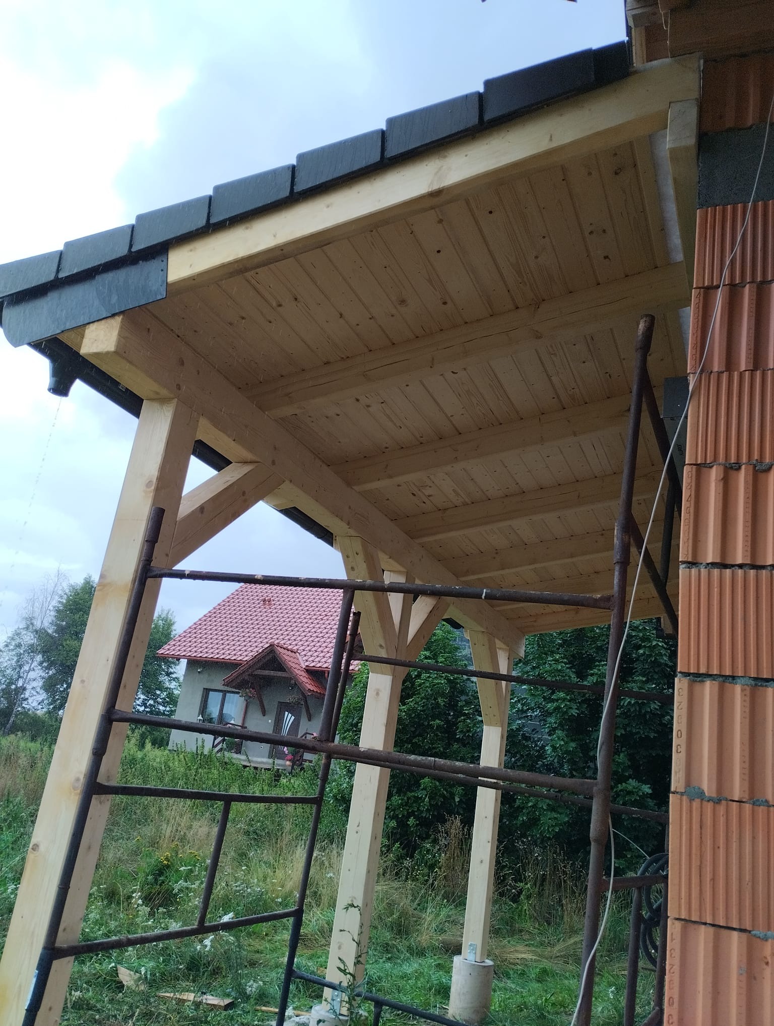 Surowa konstrukcja drewniana z czarnym dachem, w tle dom z czerwonym dachem. Widoczne rusztowanie i fragment ściany z pustaków. Realizacja w trakcie budowy.