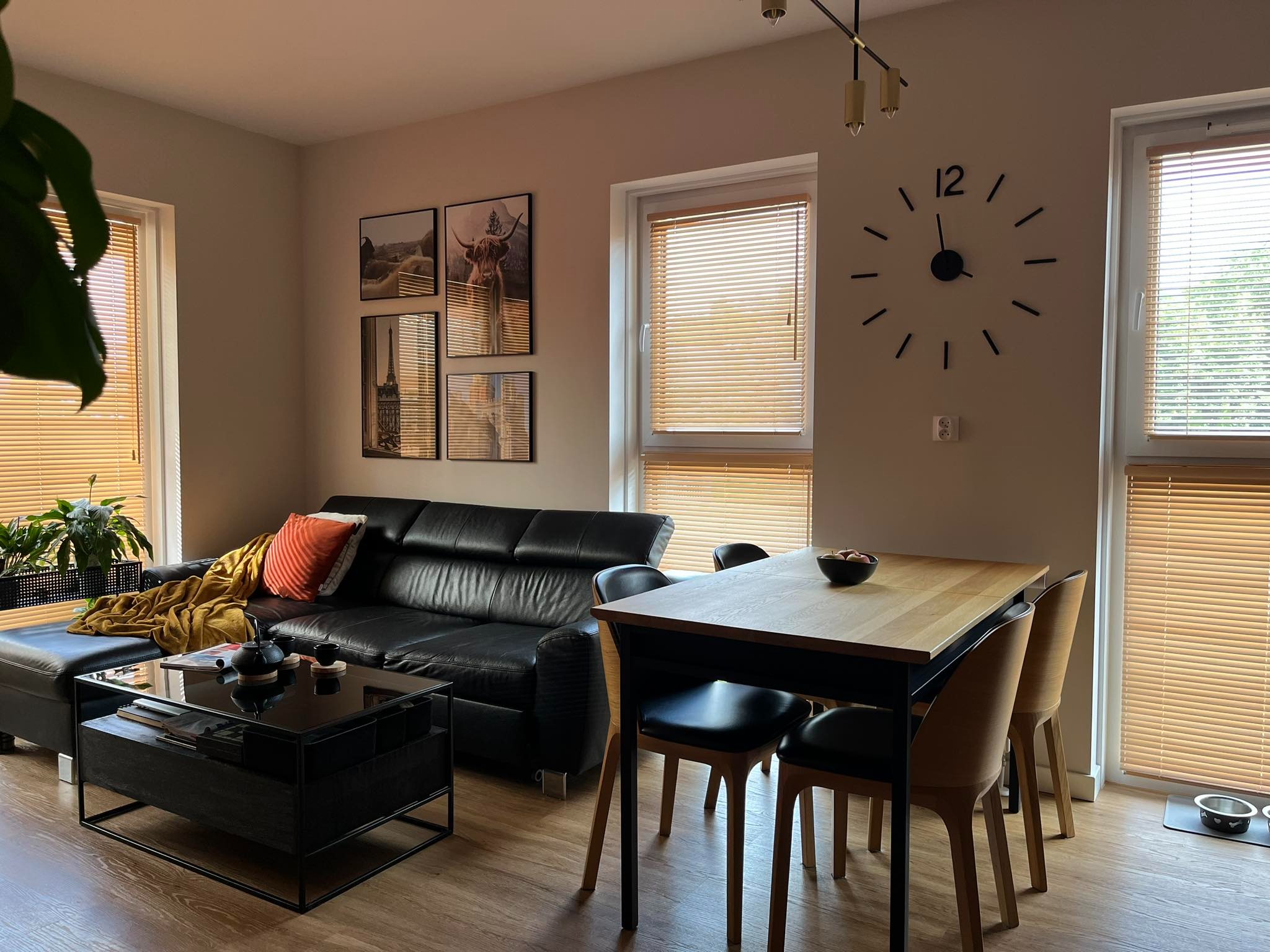 Jasne wnętrze mieszkania z czarną skórzaną sofą, stolikiem kawowym z czarną ramą i szklanym blatem, drewnianym stołem z czarnymi nogami i krzesłami oraz dekoracjami ściennymi w postaci grafik...