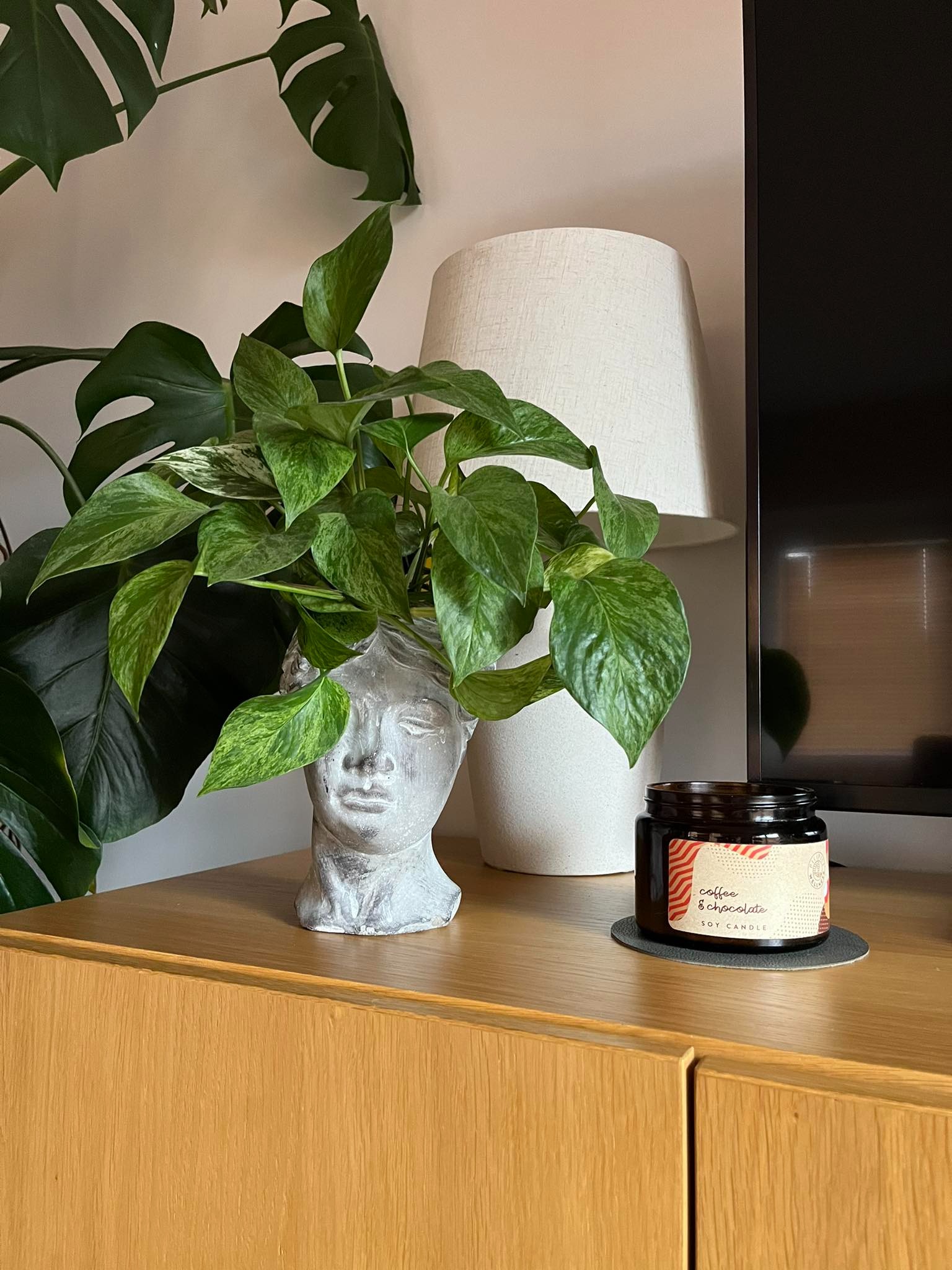 Aranżacja wnętrza w Poznaniu: roślina Pothos w popiersiu z szarego betonu na drewnianej komodzie, obok zapalona świeca sojowa 'Coffee & Chocolate' i lampa z beżowym abażurem, z widocznym fragmentem...