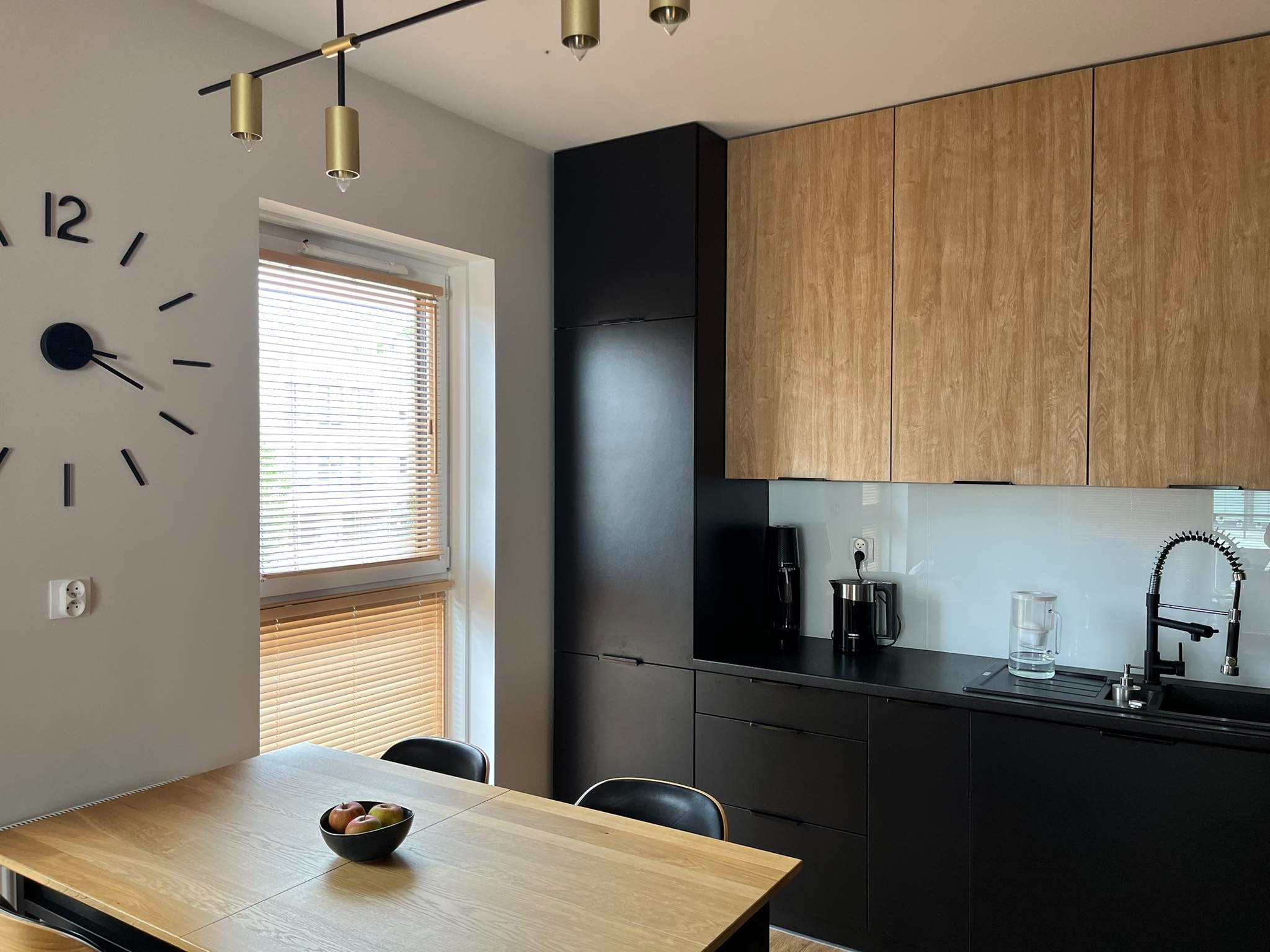 Nowoczesna kuchnia z elementami drewna i czerni. Widoczna drewniana zabudowa kuchenna, czarne szafki i blat, duży zegar na ścianie, stół z misą owoców, okno z drewnianymi żaluzjami i lampa sufitowa...