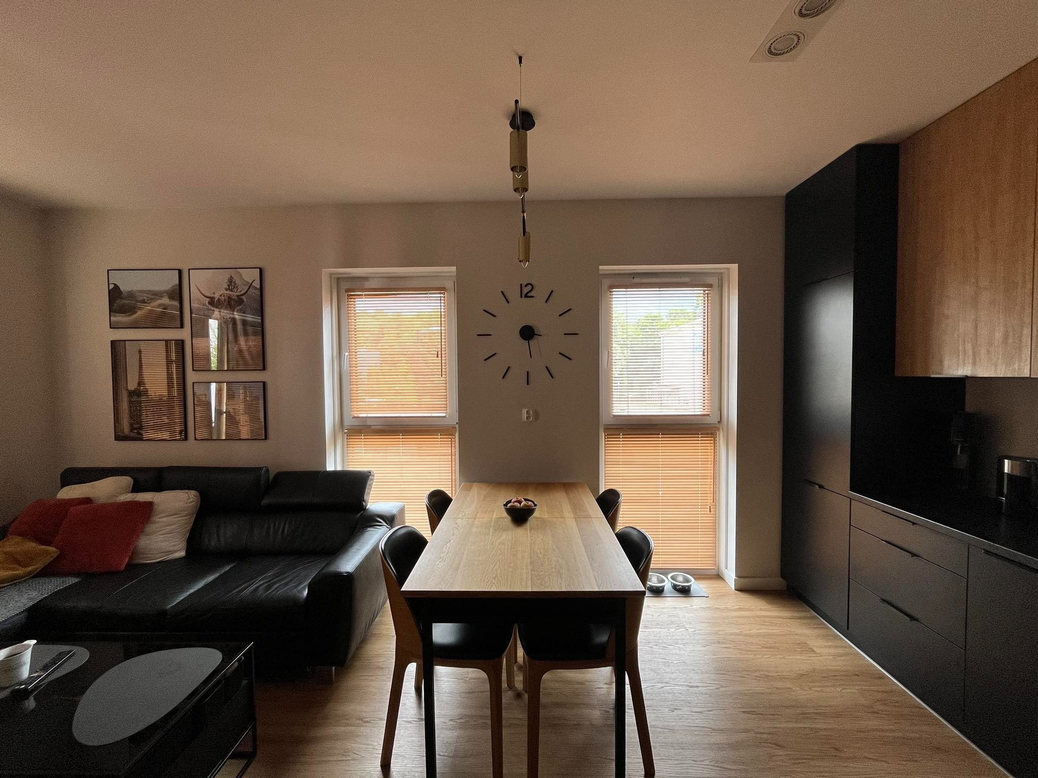 Nowoczesne wnętrze mieszkania z drewnianą podłogą, czarną skórzaną kanapą z kolorowymi poduszkami, stołem z czterema krzesłami, czarną zabudową kuchenną i zegarem na ścianie pomiędzy dwoma oknami...