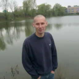 Mężczyzna w granatowej bluzie i jeansach pozuje na tle jeziora z odbijającymi się drzewami i budynkiem w tle.