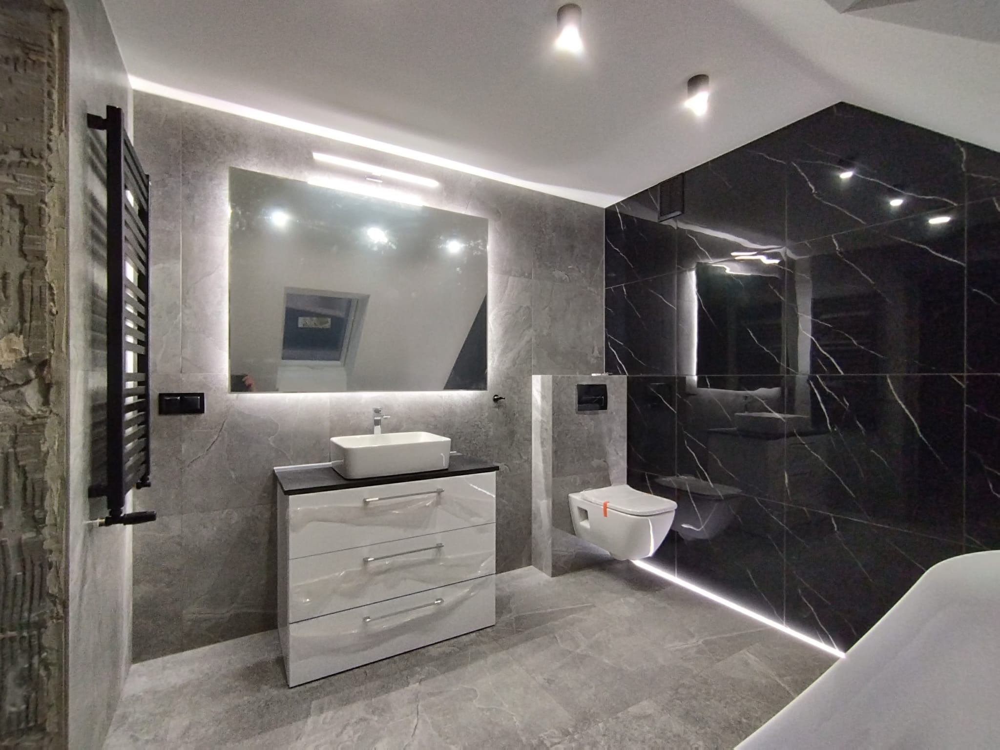 Nowoczesna łazienka z szarymi płytkami imitującymi kamień, podświetlanym lustrem, czarną ścianą z marmurowym wzorem i podwieszaną toaletą.
