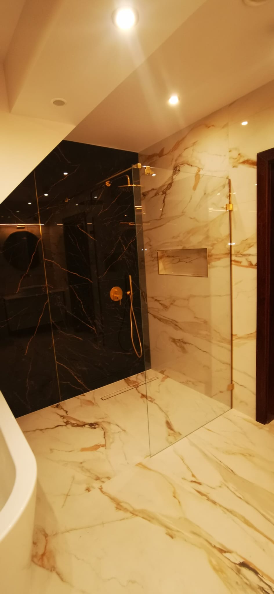 Nowoczesna kabina prysznicowa walk-in z elementami w kolorze złotym, wykończona czarnym i białym marmurem z wyraźnym użyleniem.