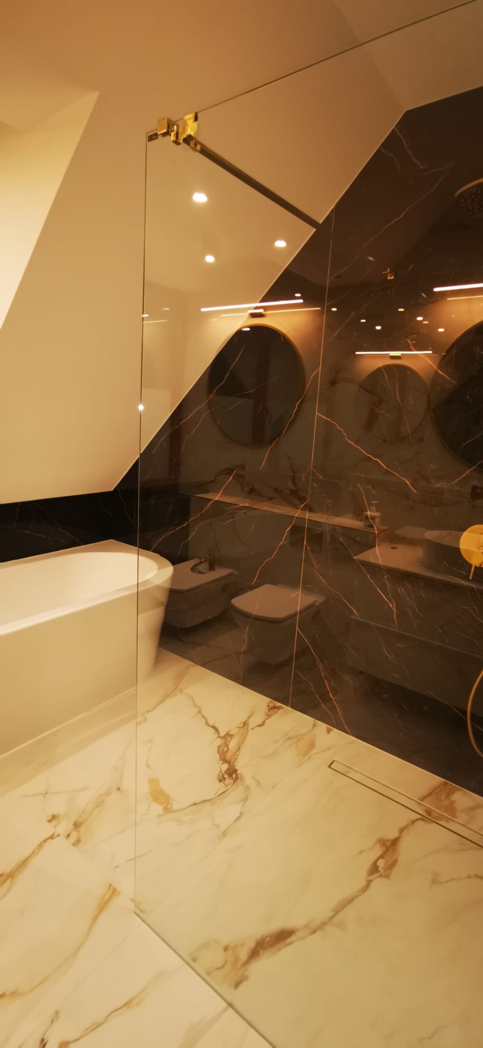 Nowoczesna łazienka na poddaszu z marmurową podłogą i ścianą w odcieniach czerni i złota, kabina prysznicowa ze złotymi okuciami, widoczne toaleta i bidet.