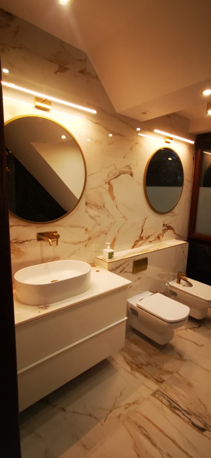 Luksusowa łazienka z marmurowymi ścianami i podłogą, okrągłymi lustrami z złotymi ramami, białą umywalką nablatową na szafce, toaletą i bidetem w stylu nowoczesnym.