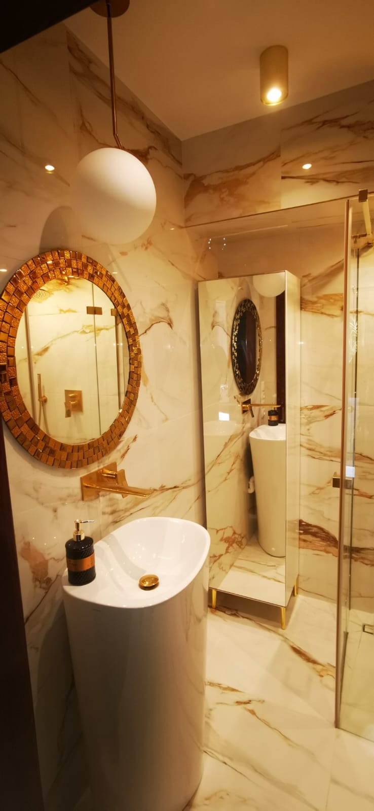 Luksusowa łazienka z umywalką na postumencie, okrągłym lustrem mozaikowym, złotą armaturą i marmurowymi płytkami ściennymi.