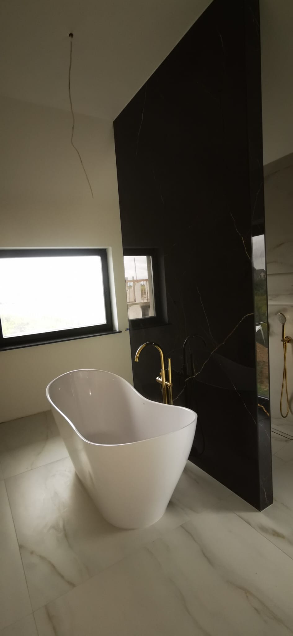 Nowoczesna łazienka z wolnostojącą białą wanną, złotą armaturą i ścianą z czarnego marmuru z złotymi żyłami, widok przez okno na budynek w budowie, białe płytki podłogowe z delikatnym wzorem.