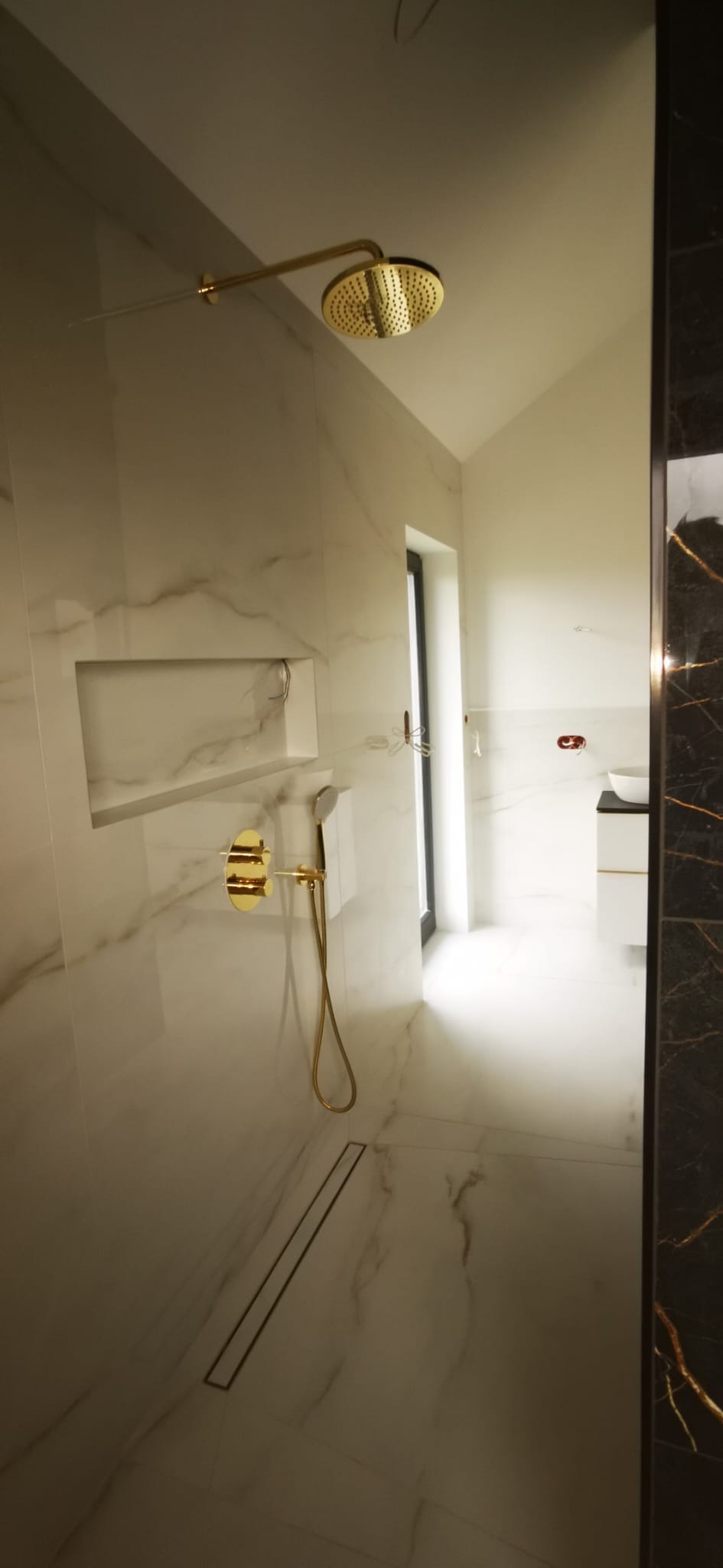 Nowoczesna łazienka z marmurowymi płytkami, złotą armaturą, odpływem liniowym i wnęką w ścianie.