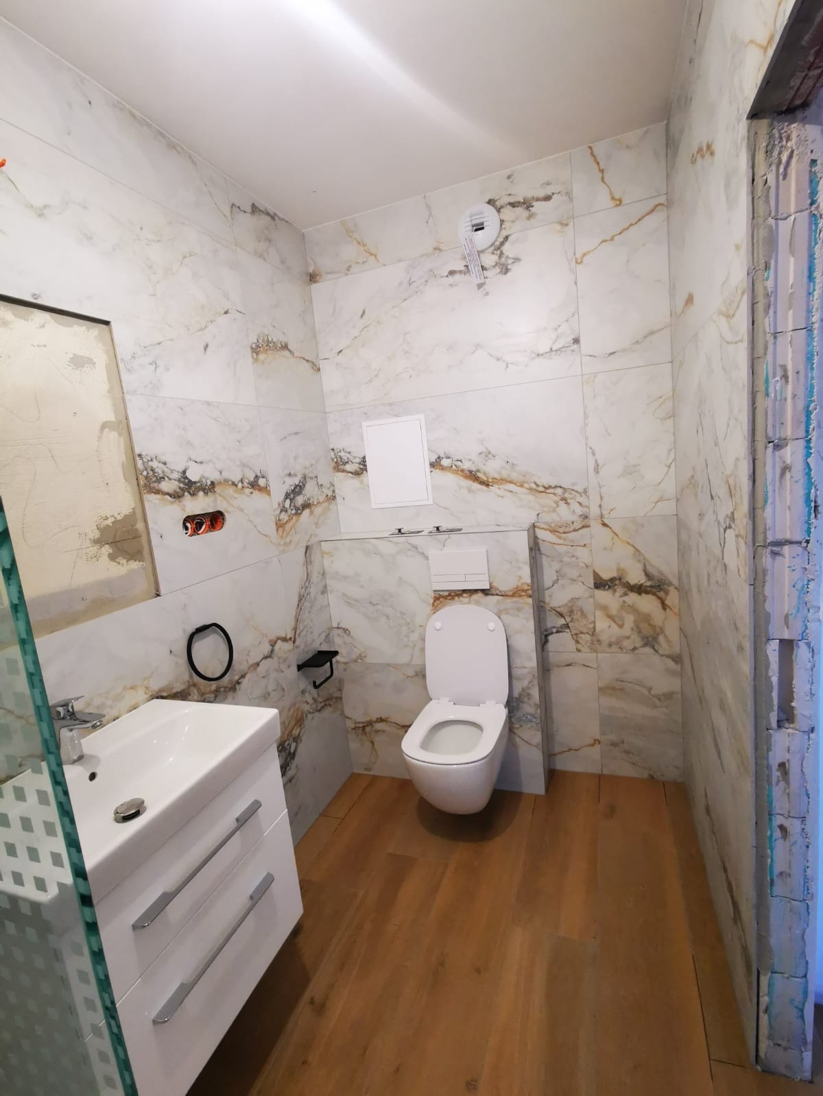 Wykończona łazienka z marmurowymi płytkami na ścianach, drewnopodobną podłogą, białą umywalką z szafką i wiszącą toaletą; widoczne niedokończone obramowanie drzwi.